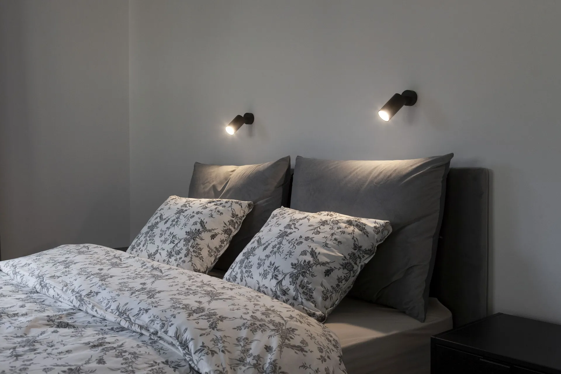 Cozy bedroom featuring a gray upholstered headboard, white floral bedding, and two black wall-mounted spotlights illuminating the space.