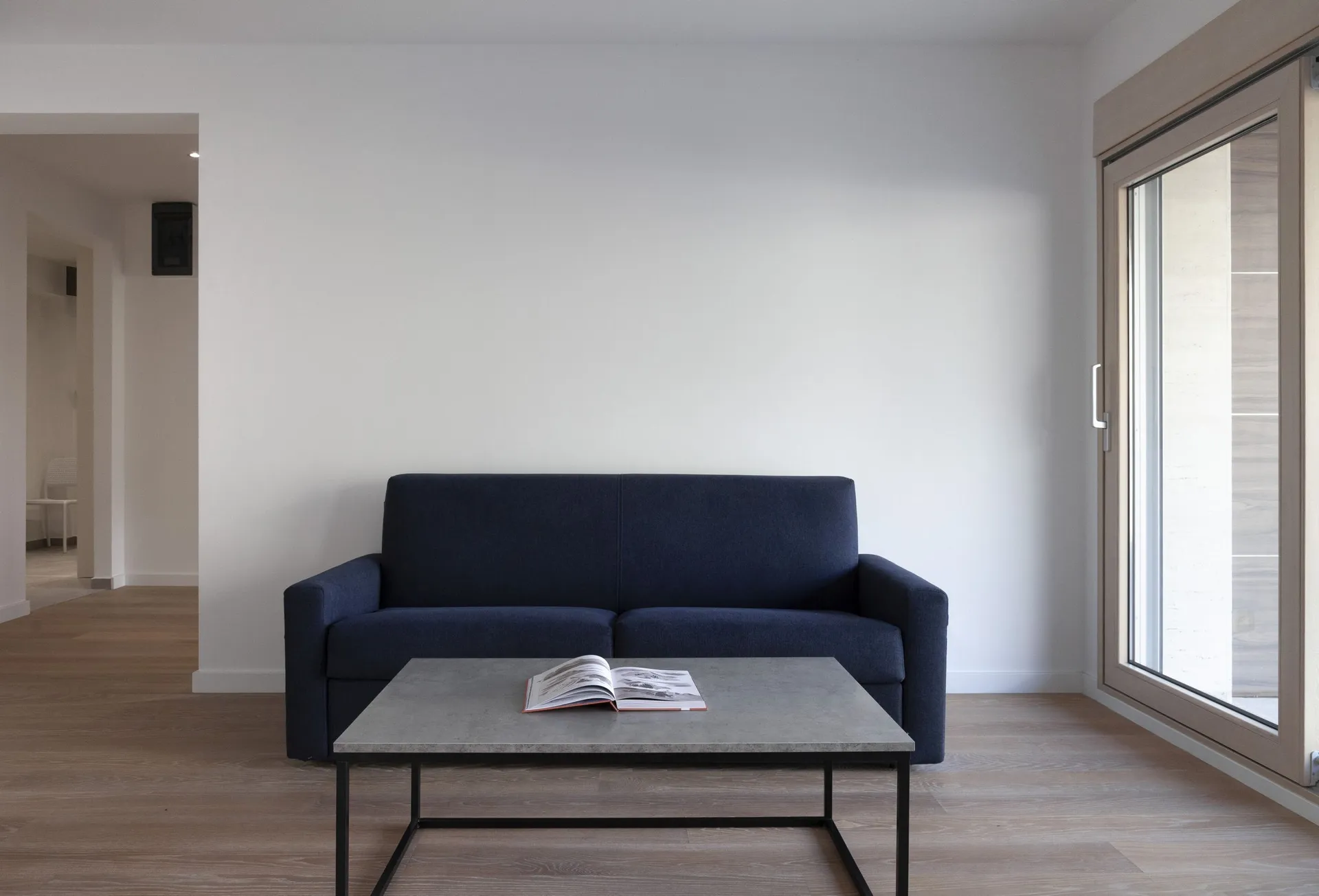 Modern living room with a dark blue sofa and grey table on light wood flooring next to a large window.