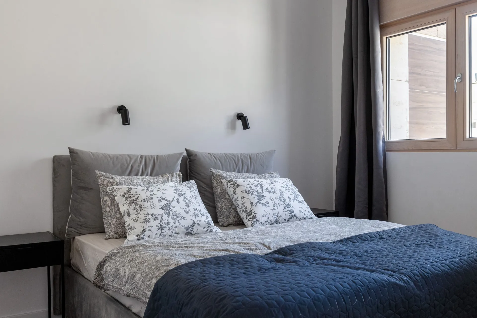Modern bedroom featuring a grey upholstered headboard, decorative pillows, and a blue quilted coverlet near the window.
