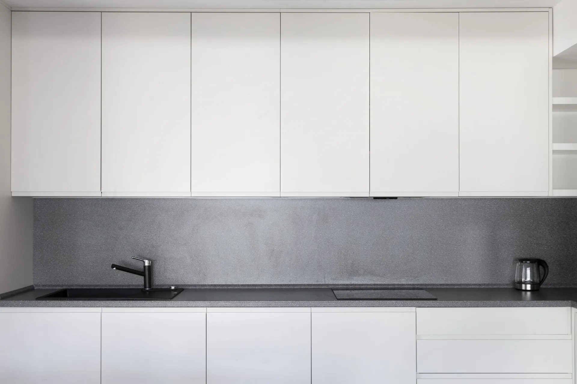 Minimalist kitchen with white cabinets, grey countertop, black sink, and glass kettle.