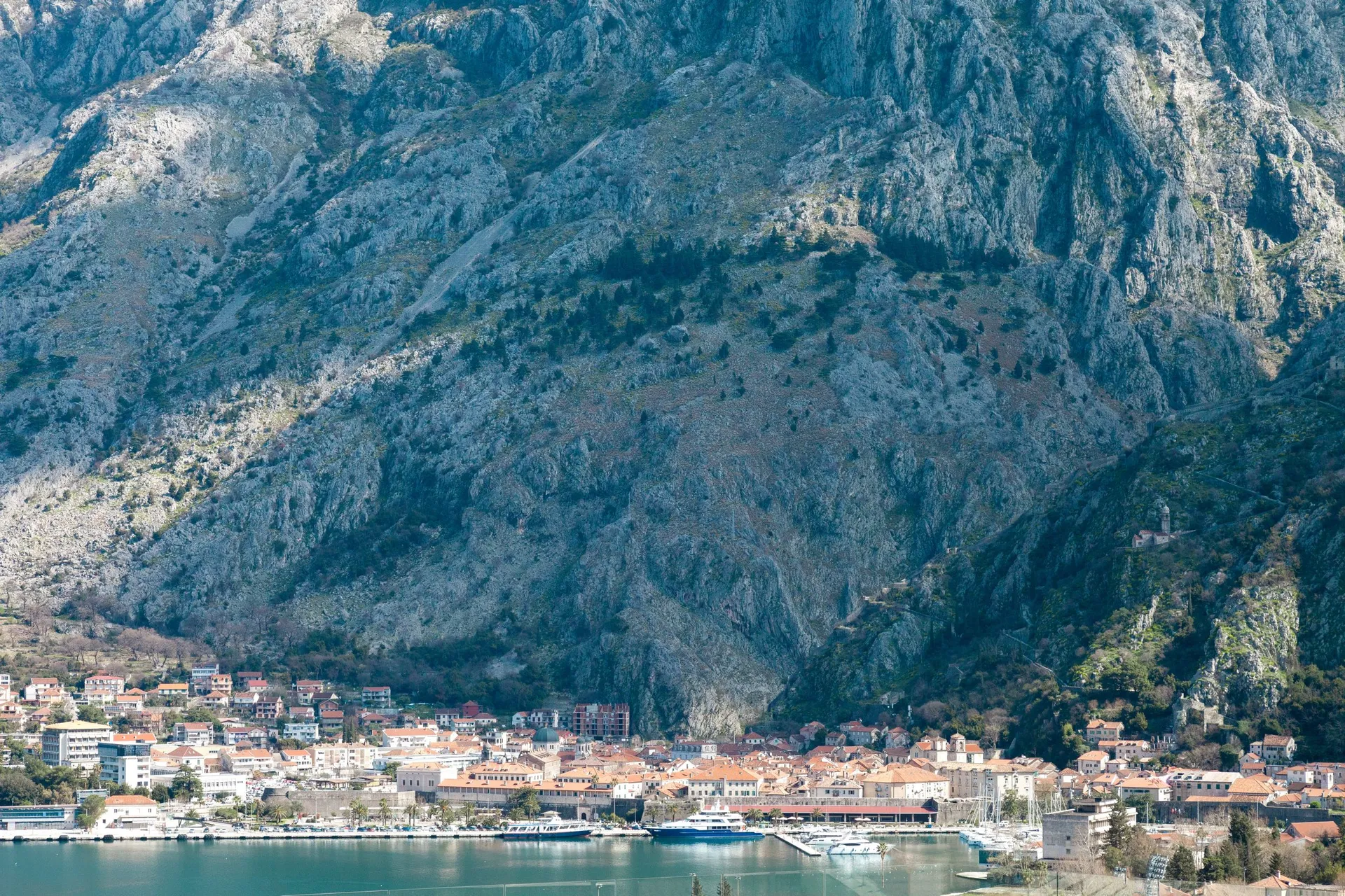 Panoramic view of a coastal town with red roofs situated at the foot of high rocky mountains and a sea harbor with boats.