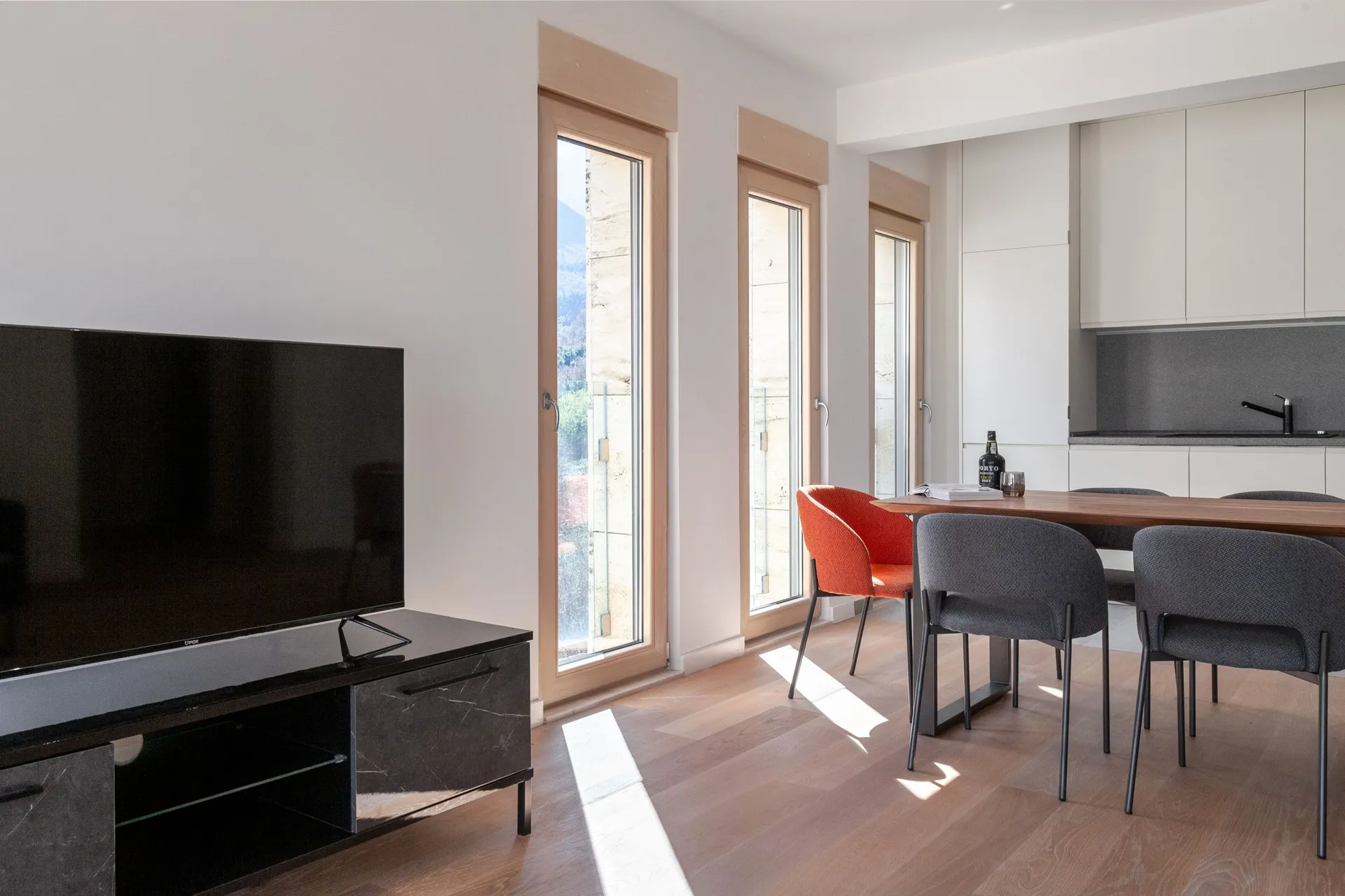 Modern living room with a TV on a black stand and a dining area next to tall windows.