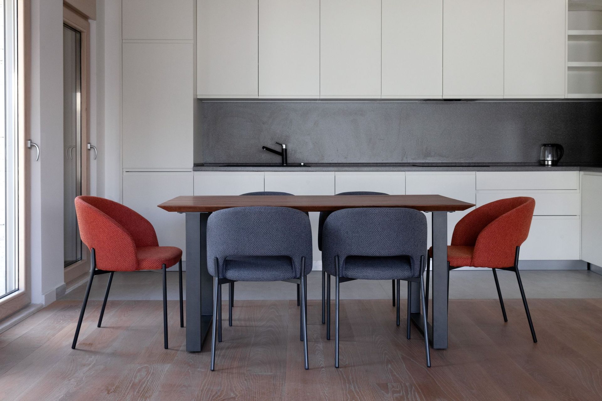 Modern kitchen in an apartment in Kotor, Montenegro: dining table, gray and red chairs, light-colored cabinetry.