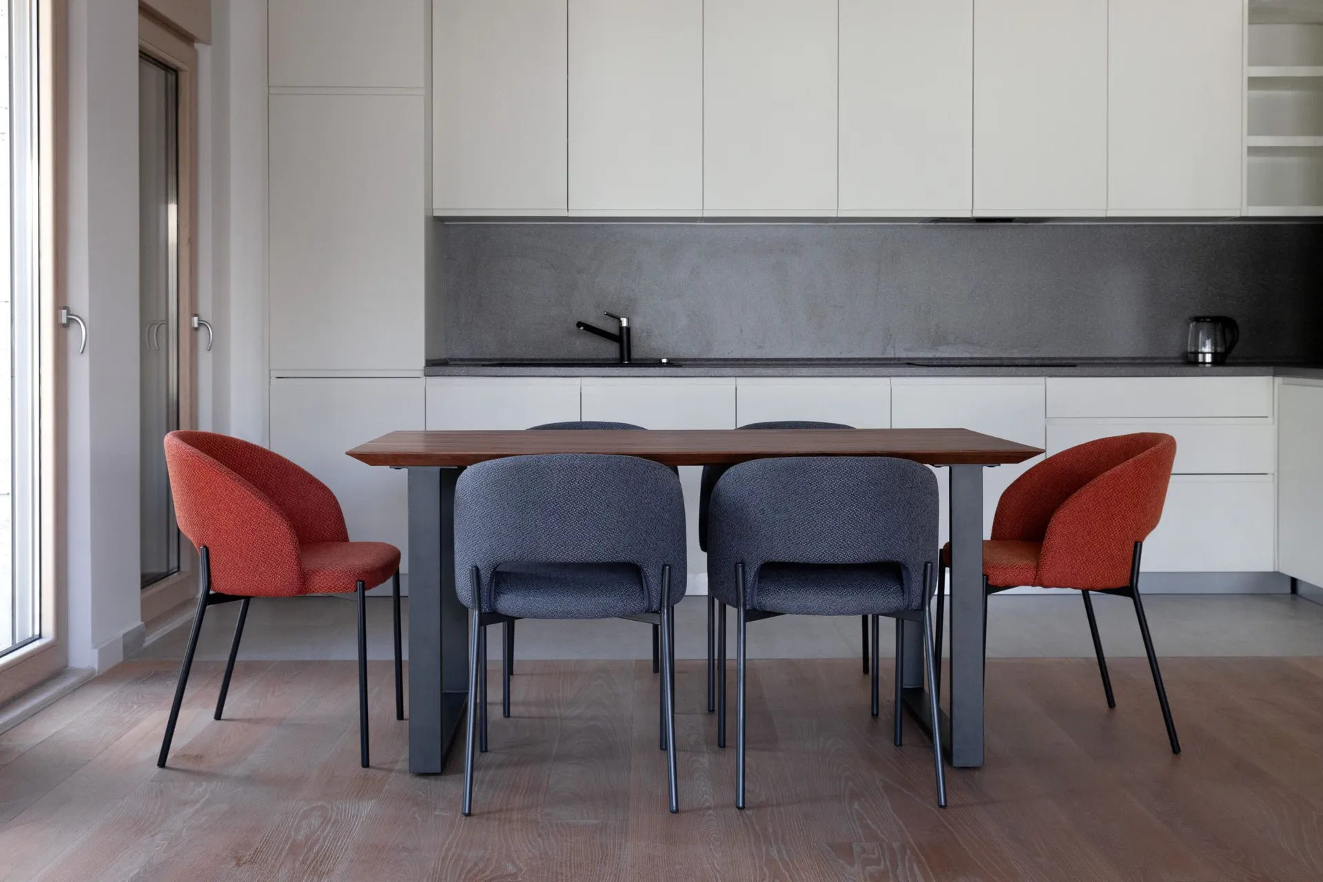 Modern kitchen with white cabinets, a wooden table, and red and grey chairs on a parquet floor.