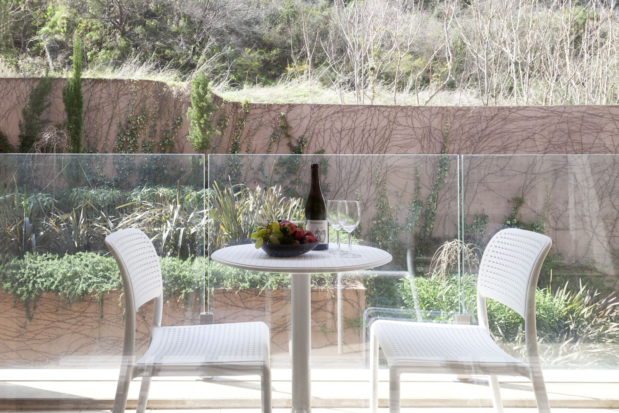 Balcony with white furniture and a view of the garden in an apartment in Kotor, Montenegro: a table with wine and fruit behind glass.