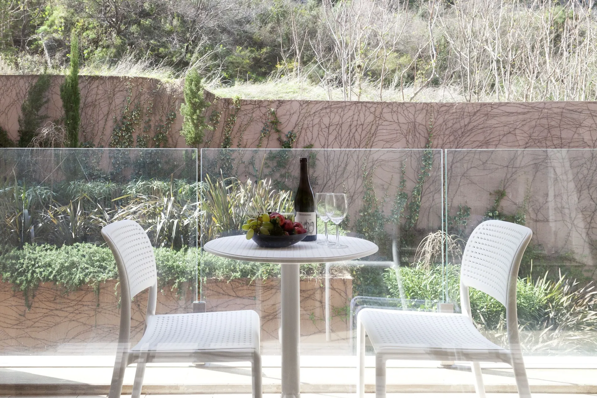 White table with two chairs, wine and fruit on a balcony with glass railing overlooking greenery.