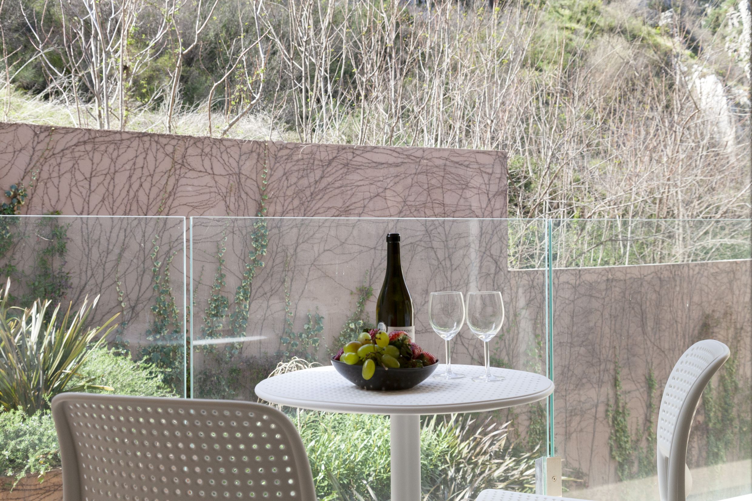 Balcony with white furniture and mountain views in apartment No. 2550, Kotor, Montenegro: a table with wine and fruit behind glass.