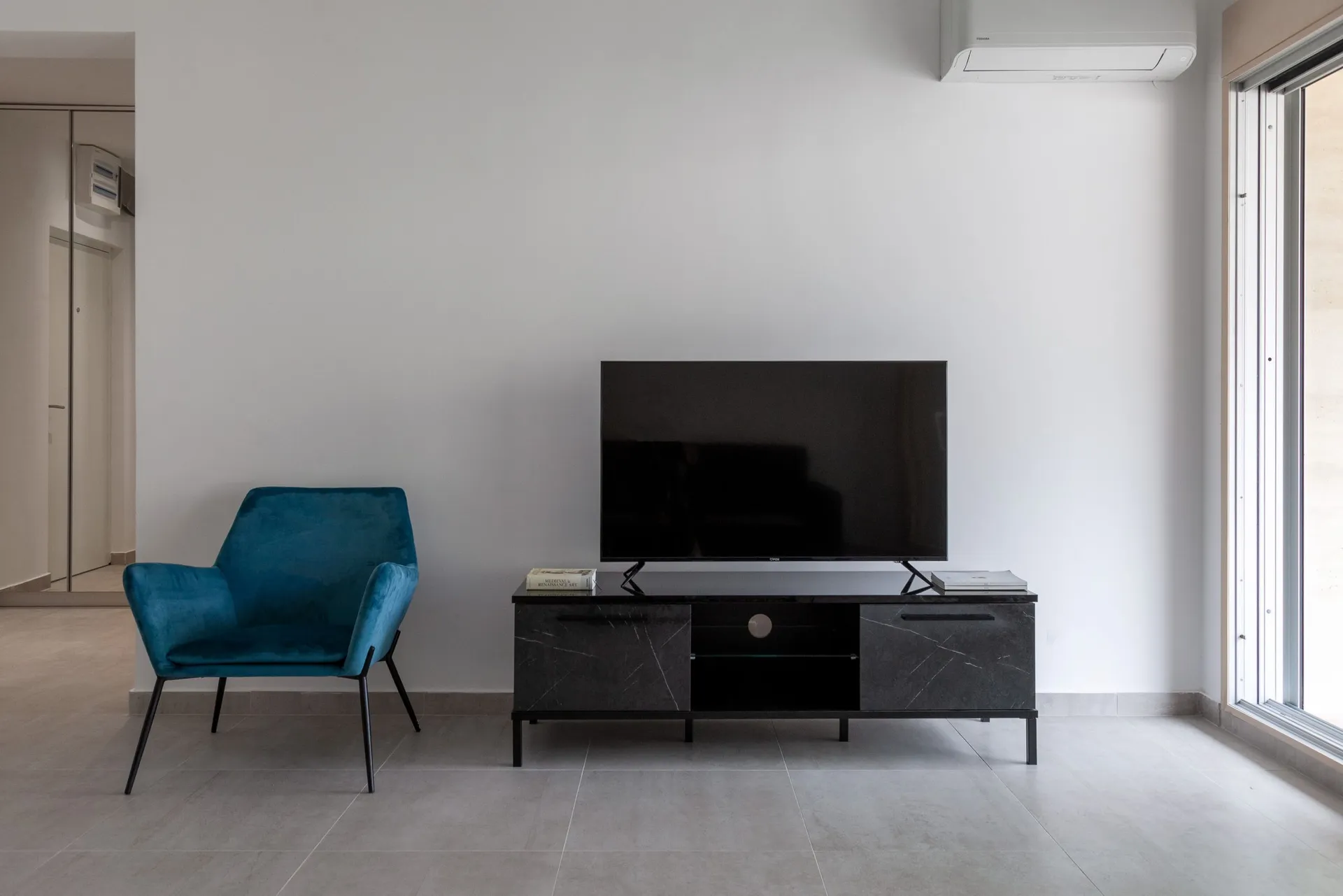Modern living room with a blue velvet armchair and TV on a black console near a bright window.