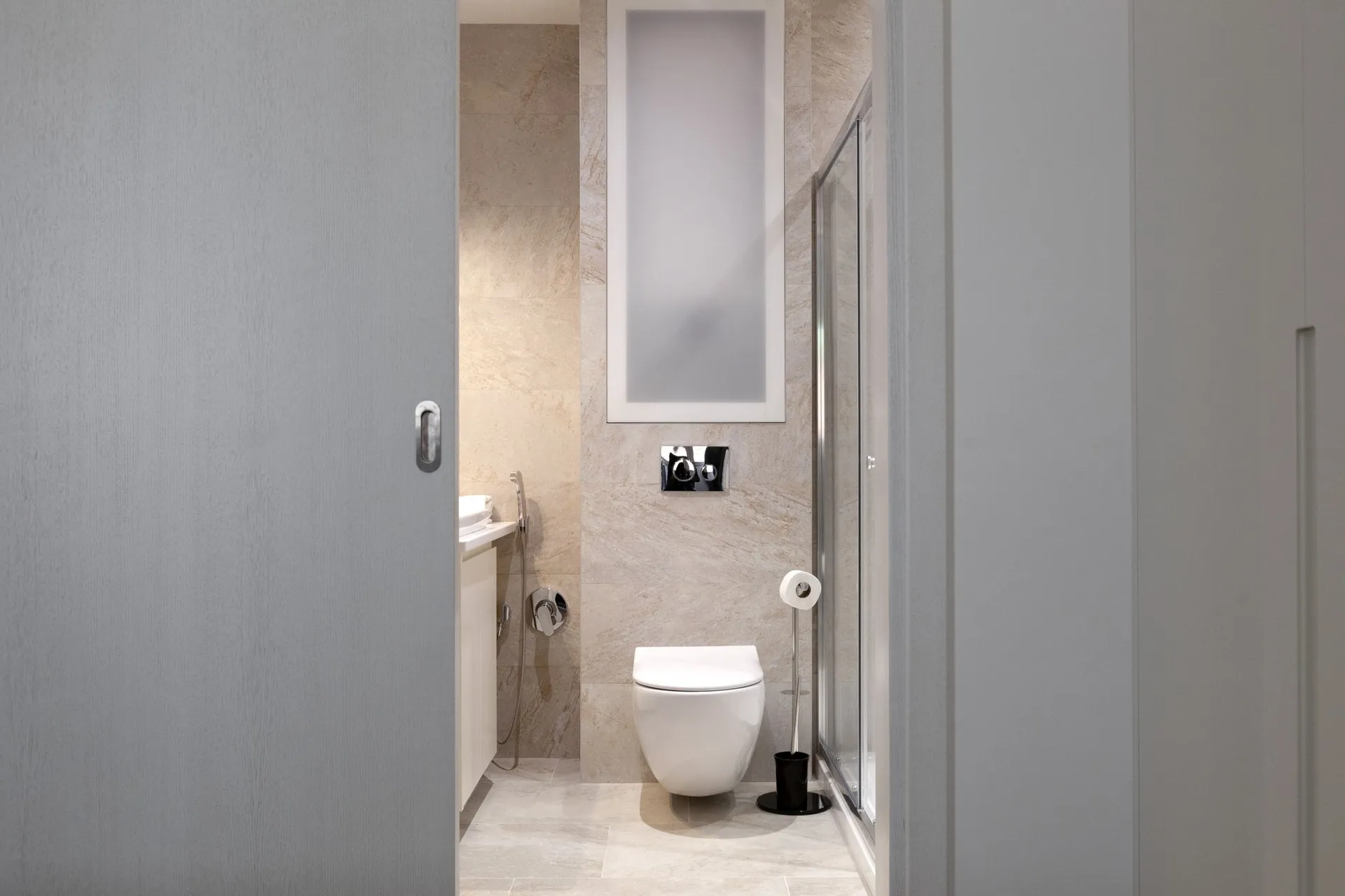 Modern bathroom featuring a wall-mounted toilet, glass shower enclosure, and beige stone tiles.