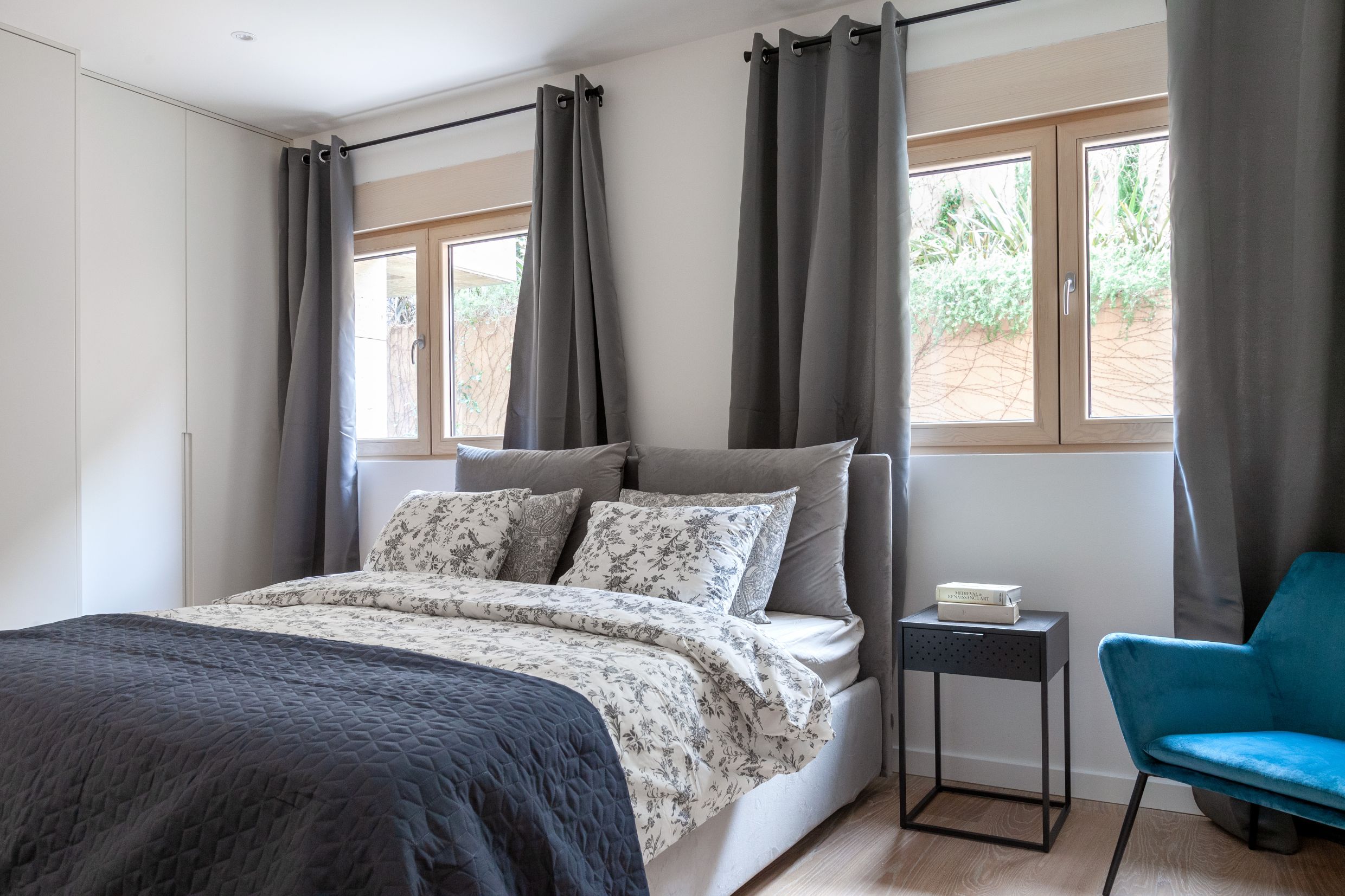 Modern bedroom in an apartment in Kotor, Montenegro: a double bed, a blue armchair, and a view of the garden from the windows.