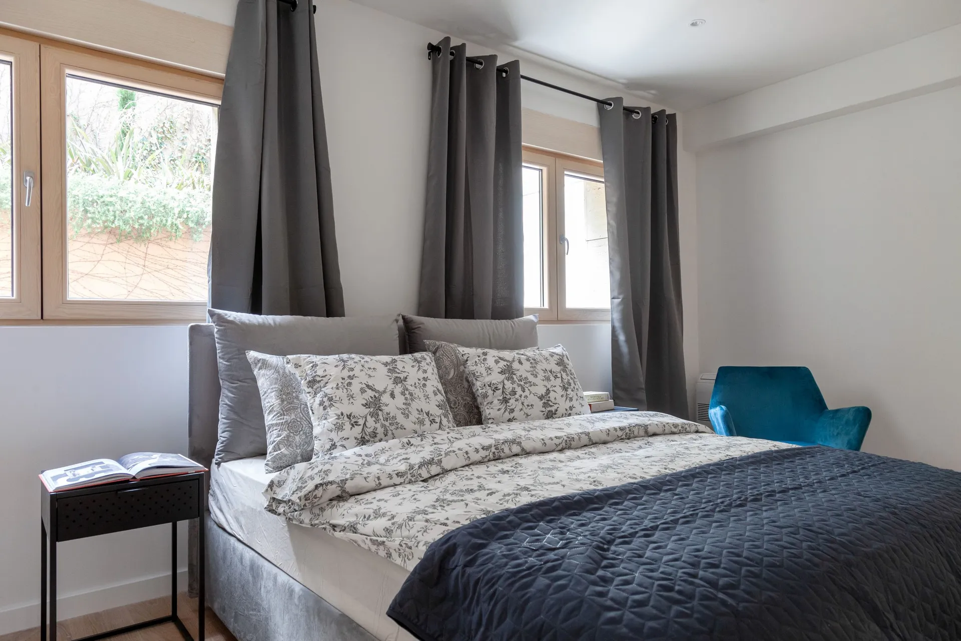 Bright bedroom featuring a double bed with floral bedding, grey curtains, and a blue chair.