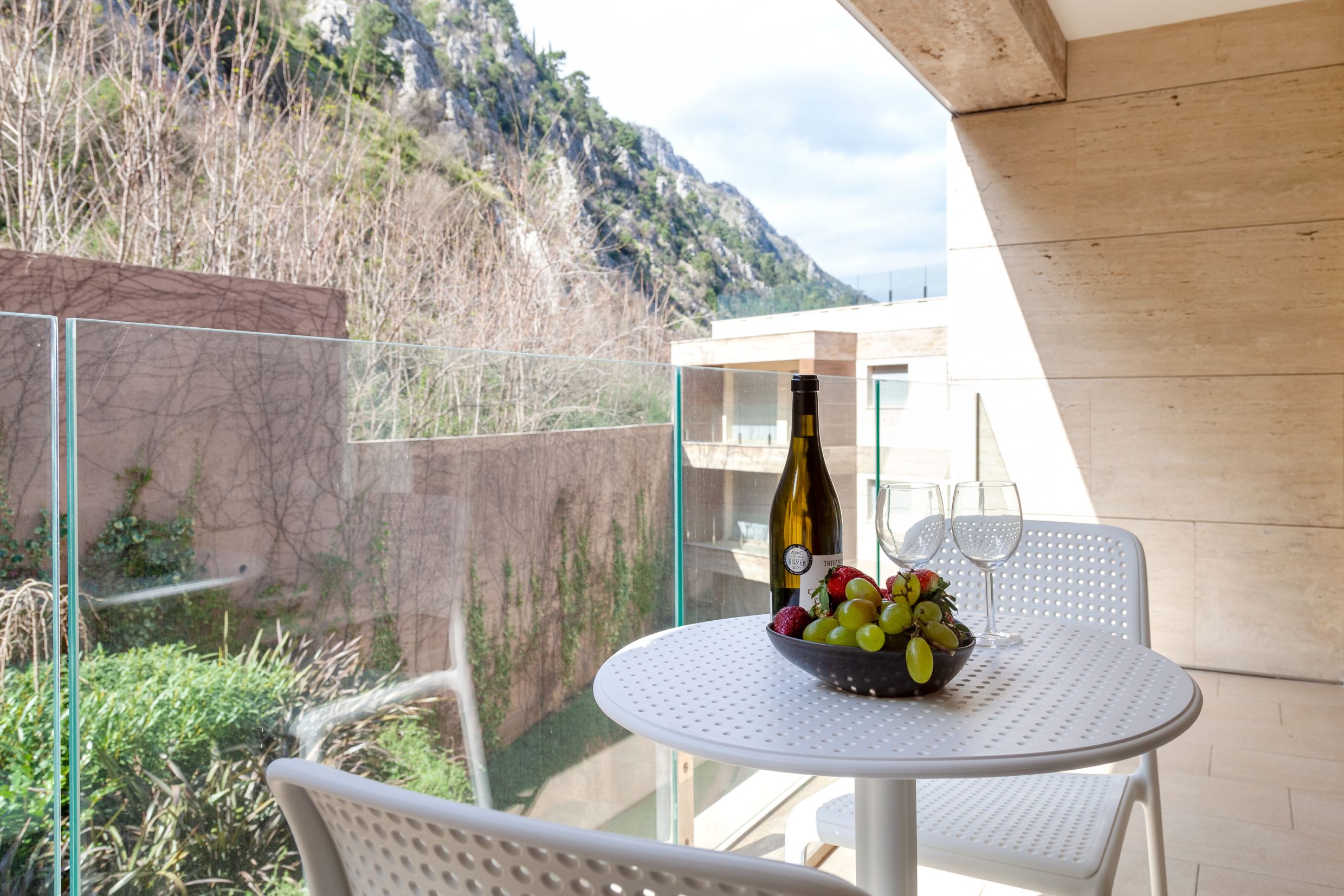 Apartment balcony in Kotor, Montenegro: a white table with wine and fruit, panoramic mountain views, and modern finishes.