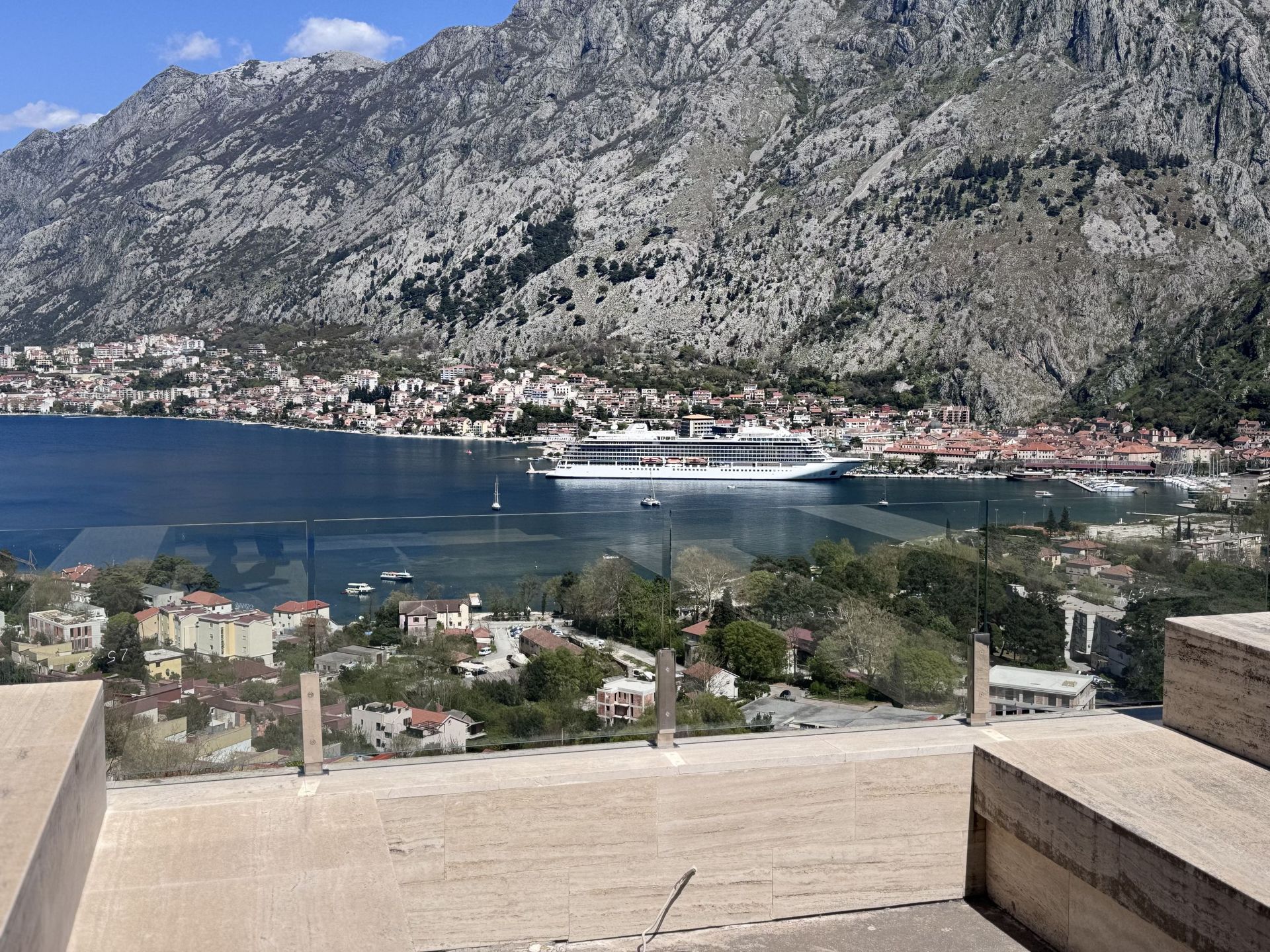 Panoramic view of the Bay of Kotor and the mountains from the apartment terrace in Kotor, Montenegro, with a glass railing.
