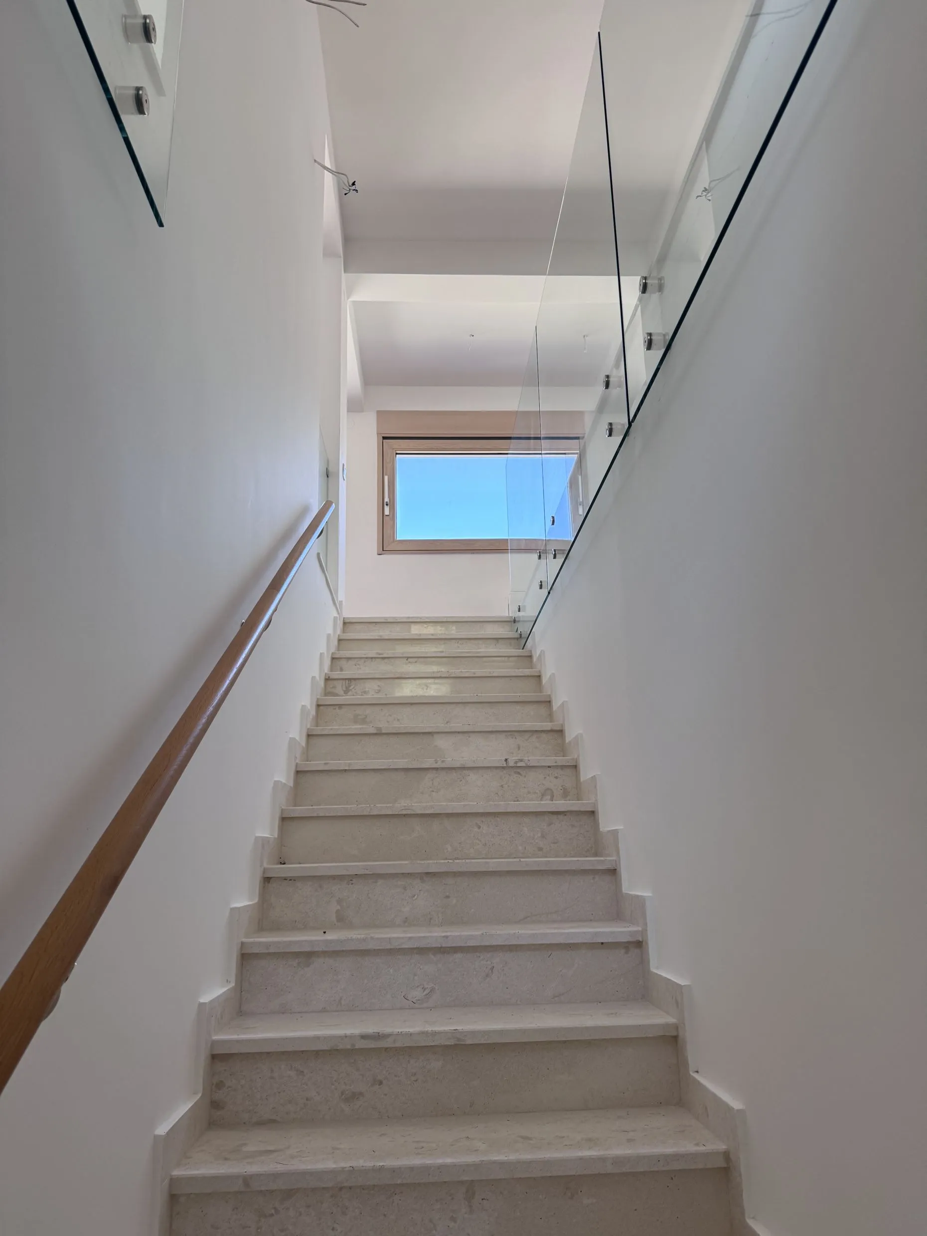 Bright staircase with light stone steps, a wooden handrail, and a glass balustrade leading to a window.