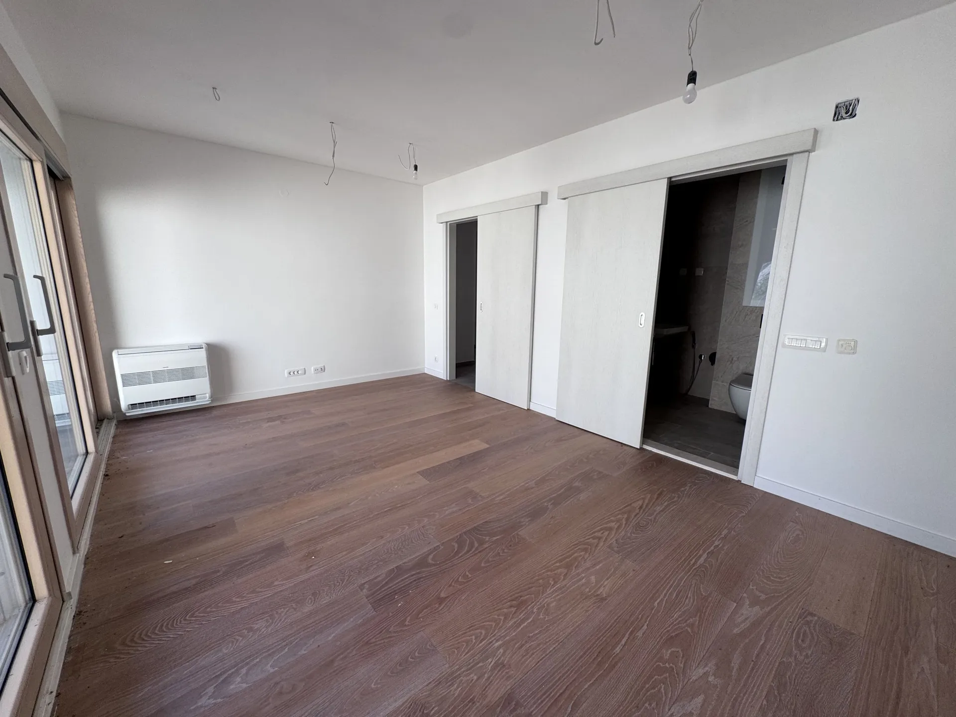 Empty room with wooden flooring, white walls, wall-mounted AC unit, and sliding doors leading to a bathroom.