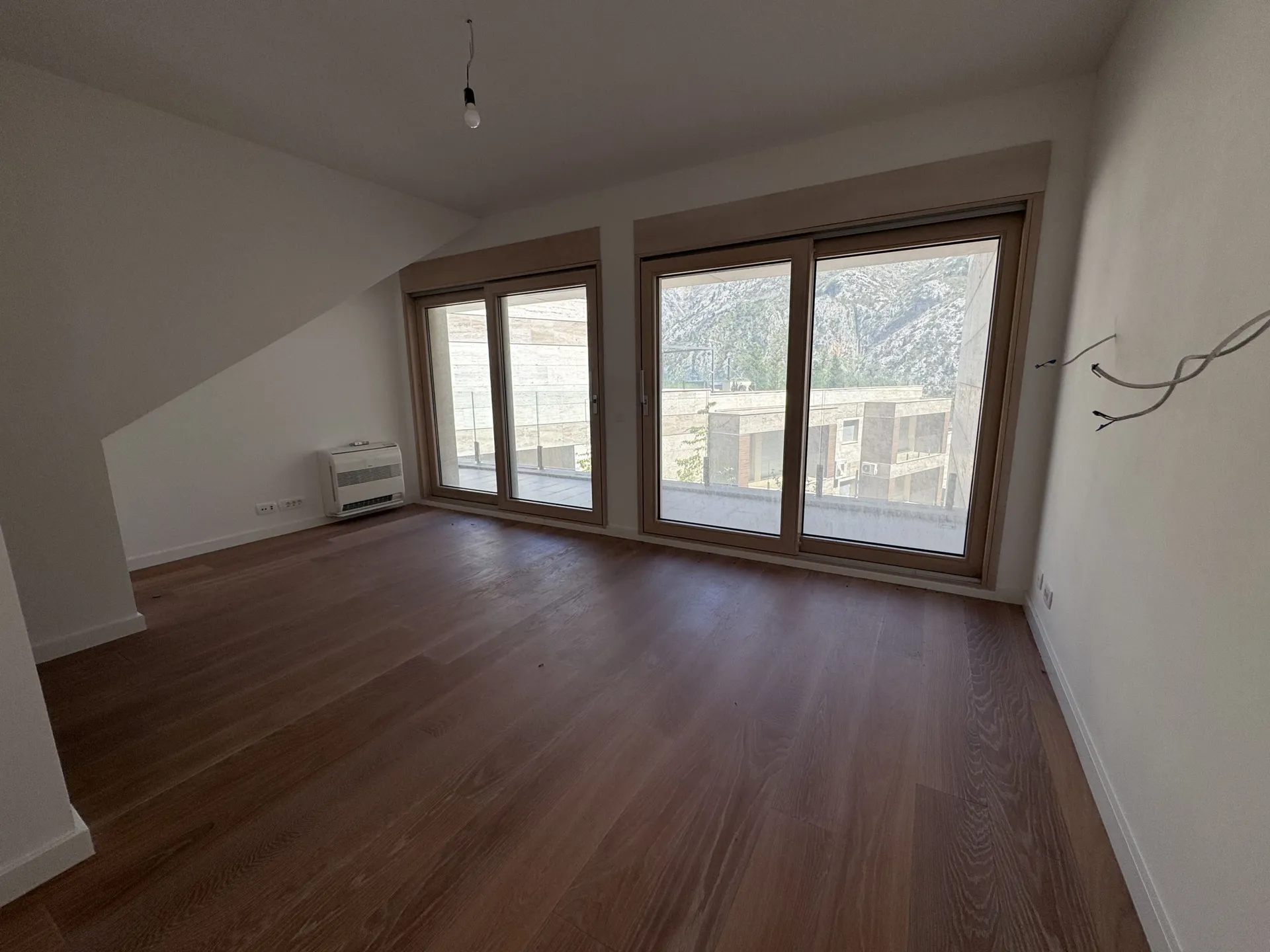 Empty room with dark wood flooring and large windows overlooking mountains. A white climate unit and exposed wires are visible on the walls.