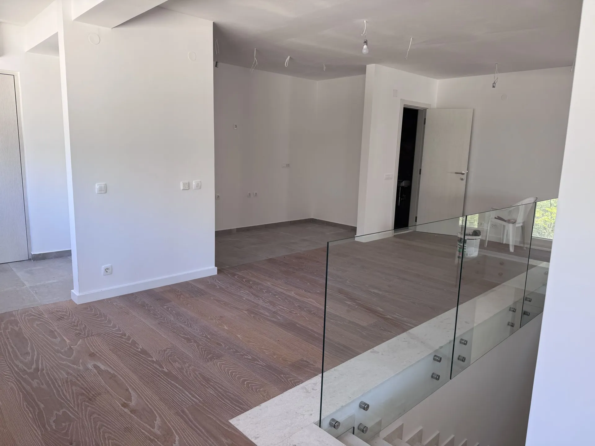Empty room with wooden flooring and glass staircase railing. White walls, hanging light bulbs, and an open door visible.