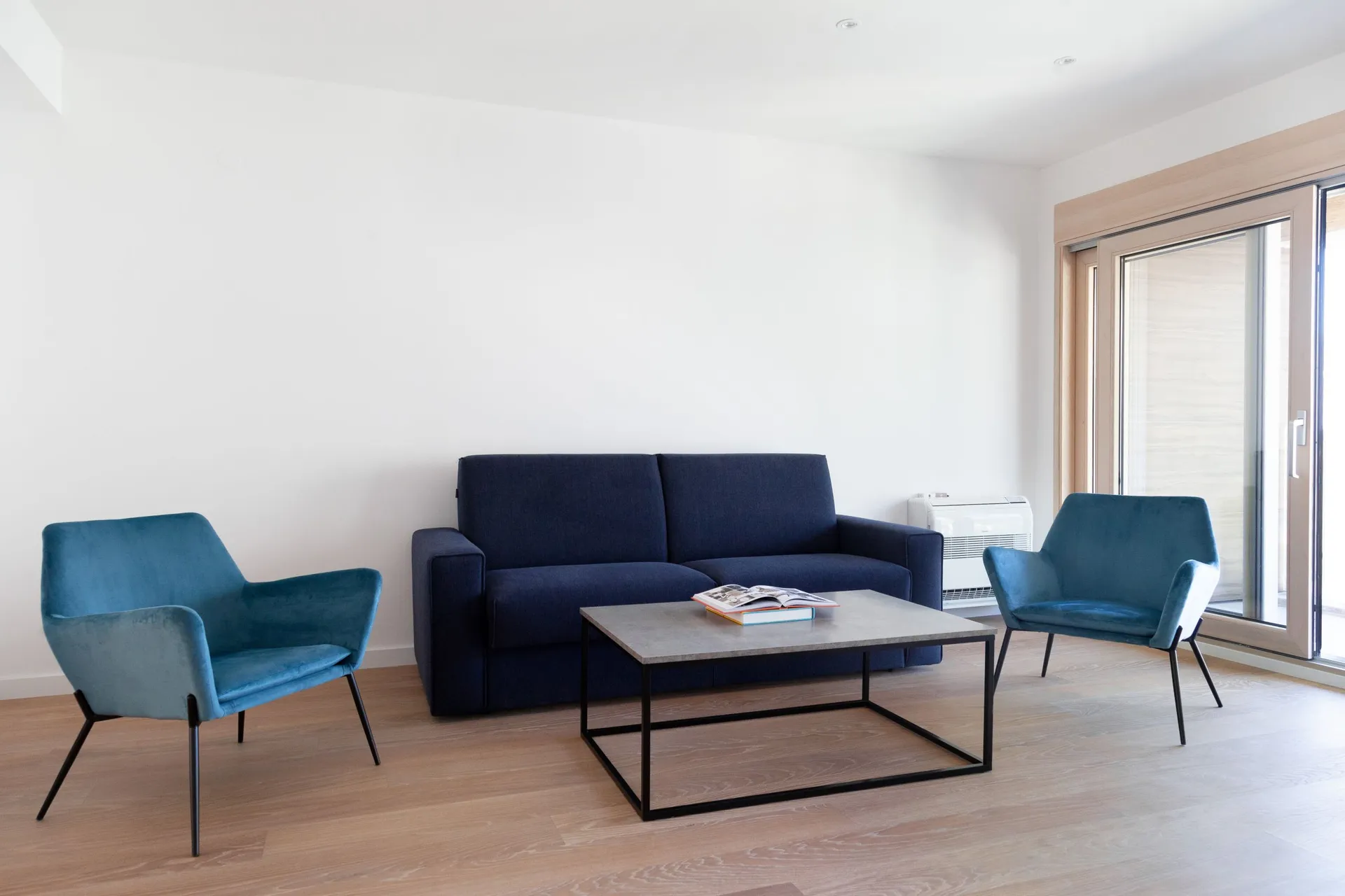 Modern living room featuring a dark blue sofa, two teal armchairs, and a grey coffee table on a black frame near a window.