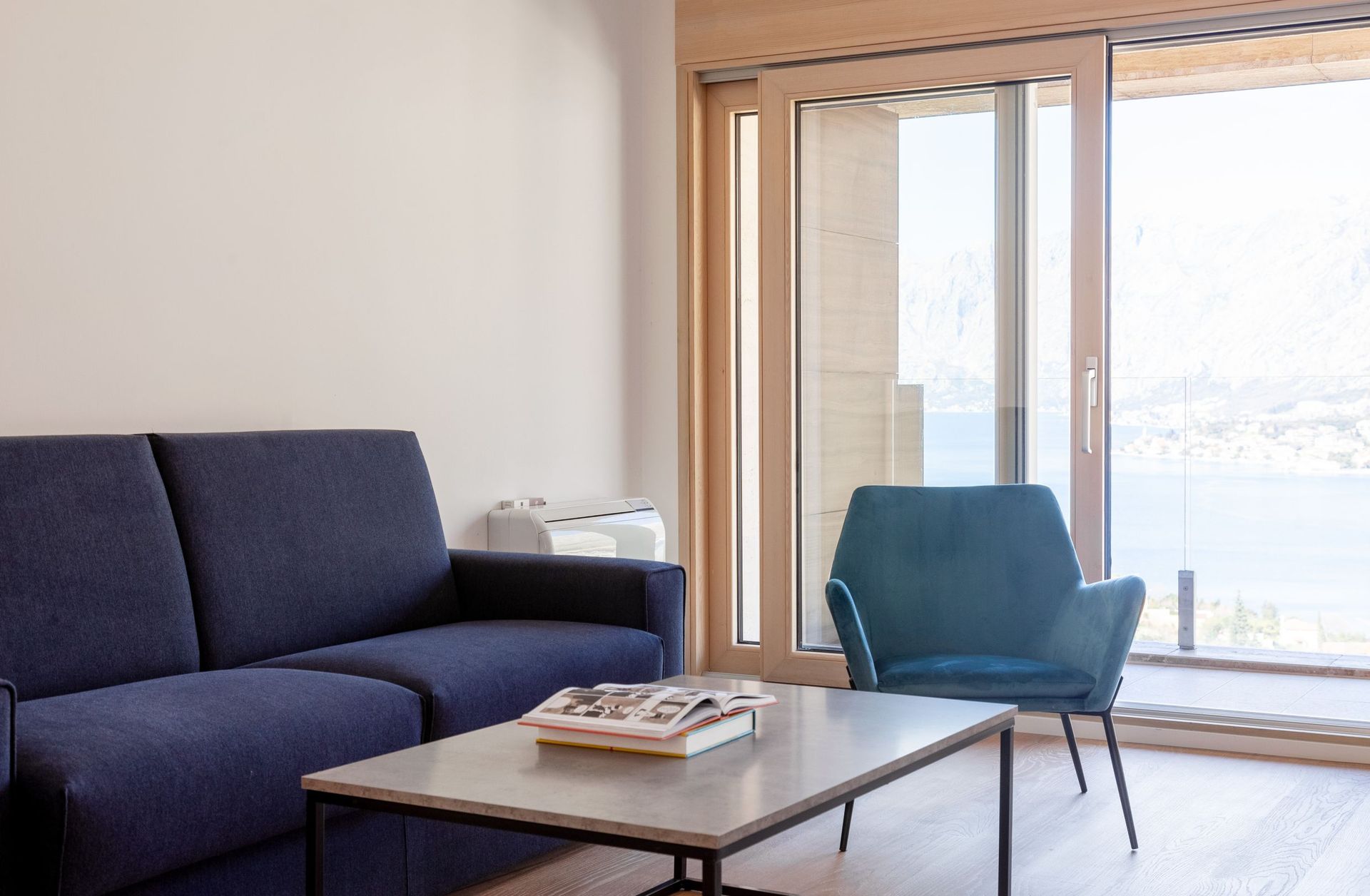 Living room with a blue sofa and armchair in an apartment in Kotor, Montenegro, with panoramic sea and mountain views through the balcony.