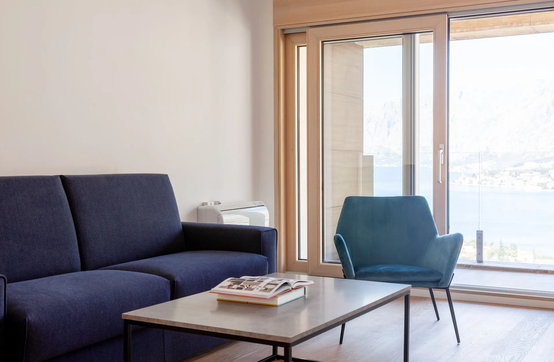Modern living room with a blue sofa, armchair, and coffee table. Large window offers a panoramic view of the sea and mountains.