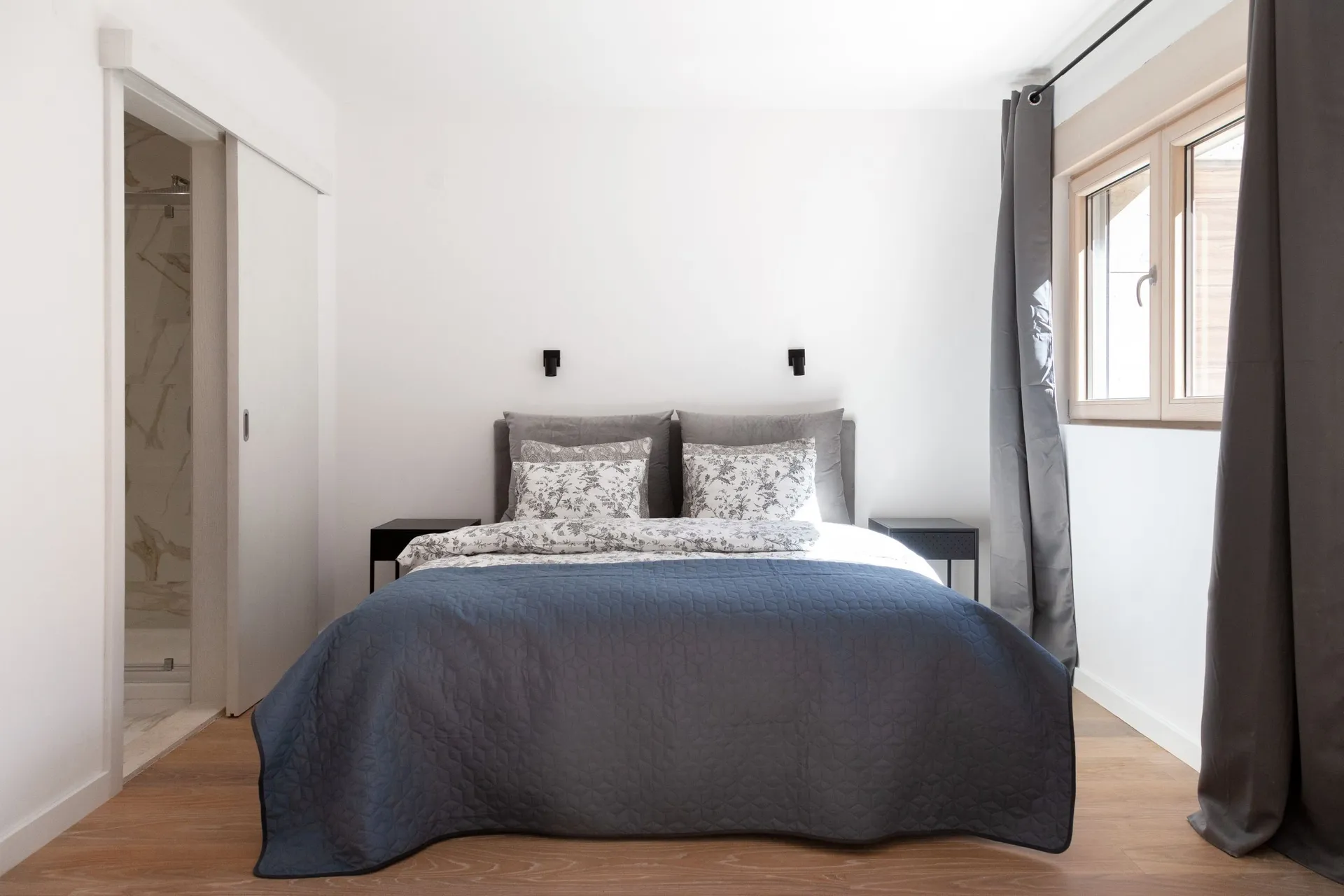 Modern bedroom with a double bed, grey quilt, and floral pillows. An open door to the bathroom is visible on the left.