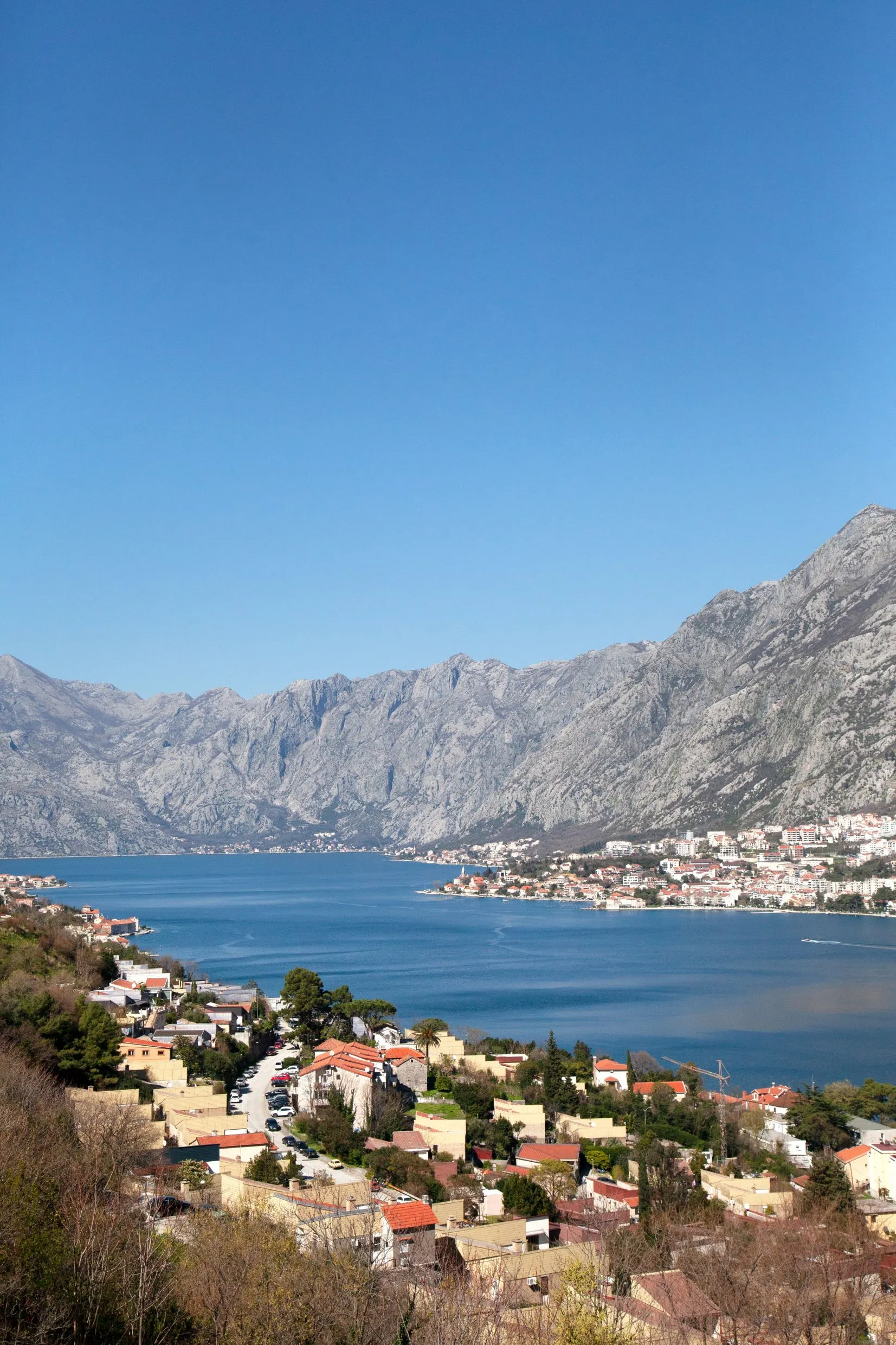 Panoramic view of a blue sea bay surrounded by high rocky mountains and a town with red roofs under a clear blue sky.