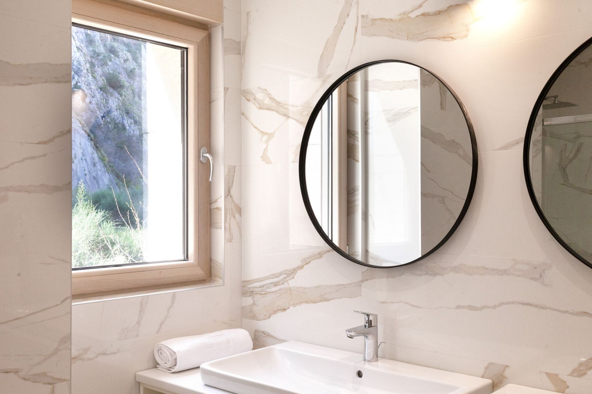 Modern bathroom with marble-style finishes in an apartment in Kotor, Montenegro, with a round mirror and mountain view.