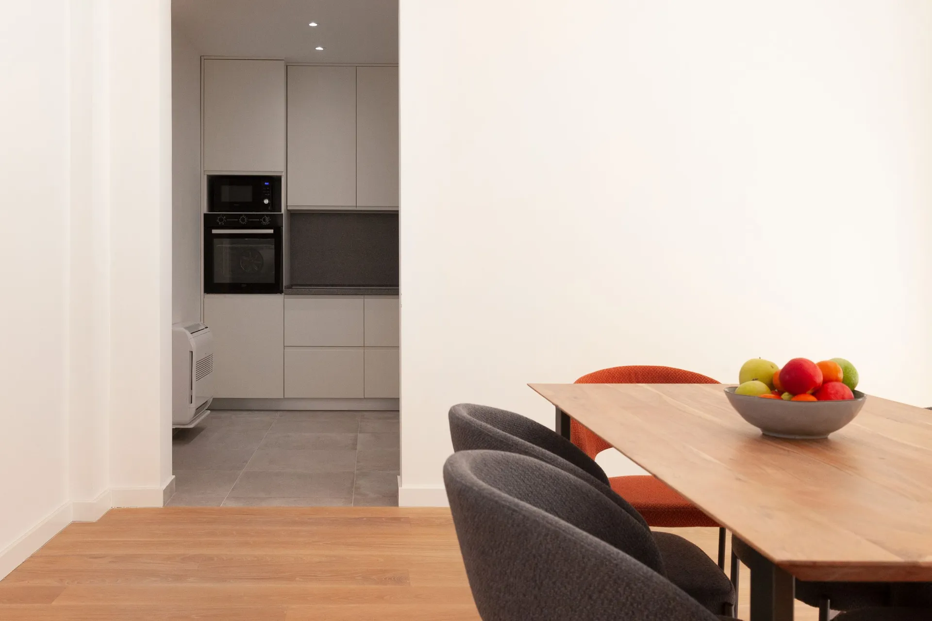 Modern kitchen with grey cabinets and built-in appliances. A wooden table with a bowl of fruit and upholstered chairs is in the foreground.