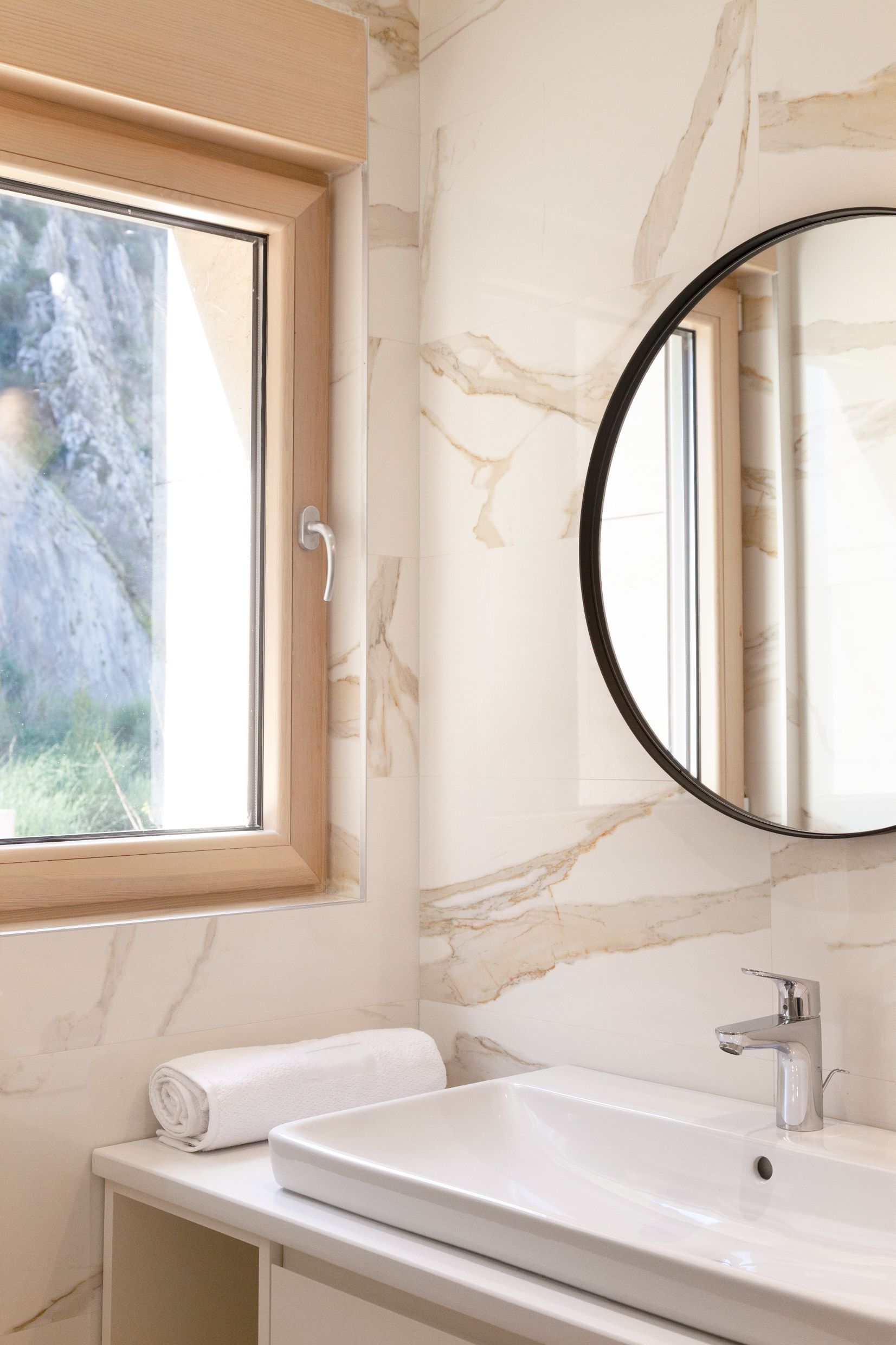 Modern bathroom in an apartment in Kotor, Montenegro: light marble-look tile, round mirror, and mountain view.
