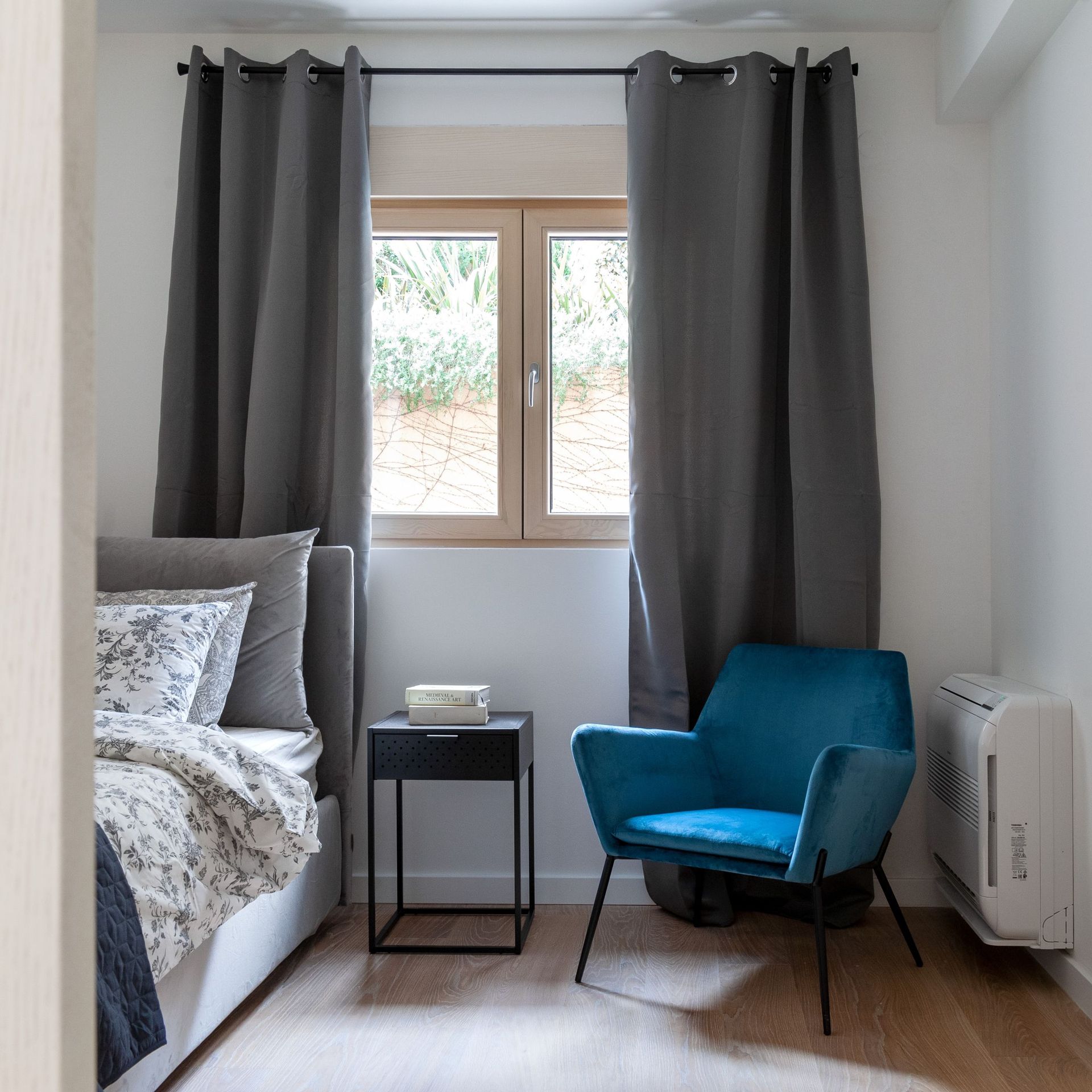 Cozy bedroom in an apartment in Kotor, Montenegro: a gray bed, a blue velvet armchair, and a window view of greenery.