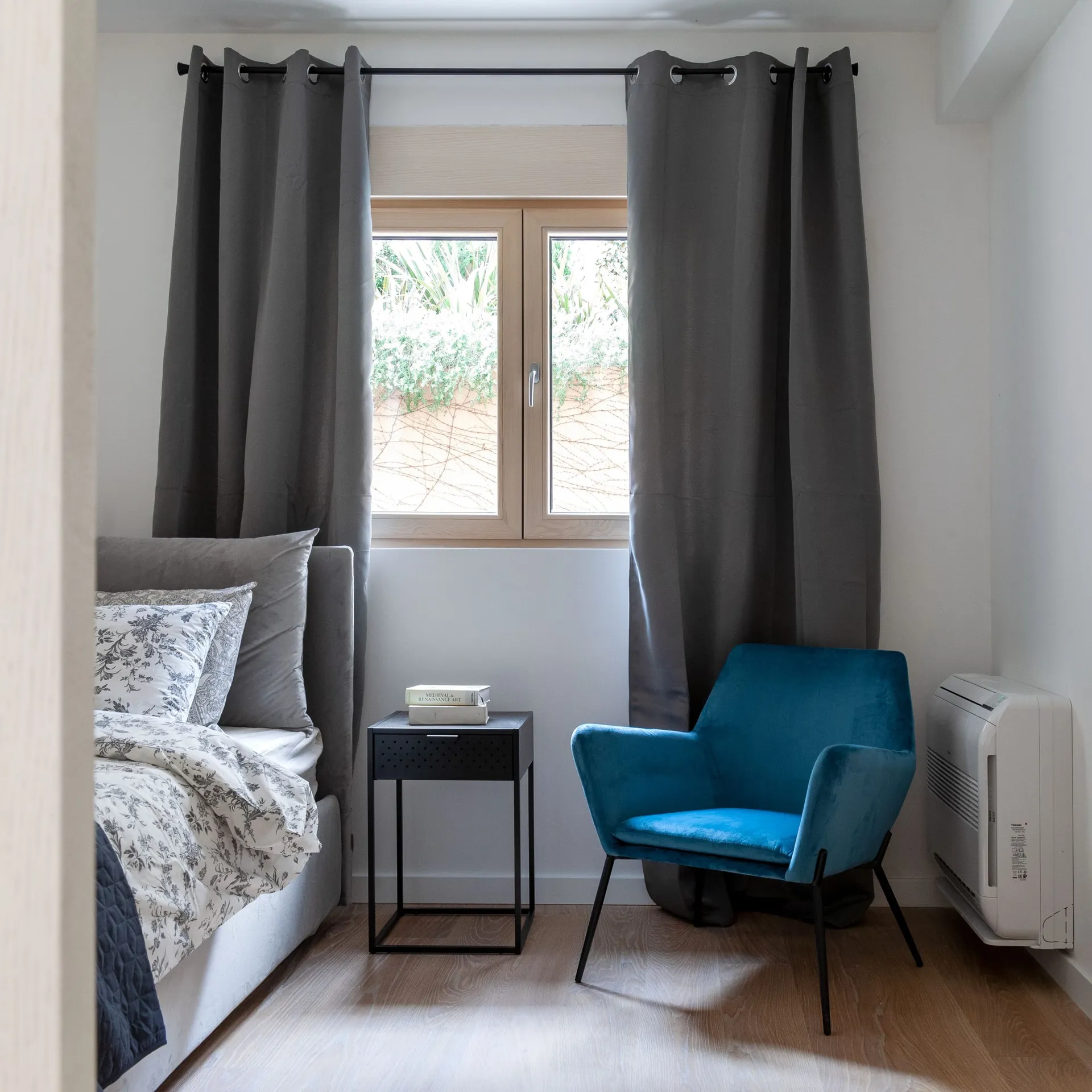 Modern bedroom featuring grey curtains, a blue velvet armchair, and a bed with floral bedding near the window.