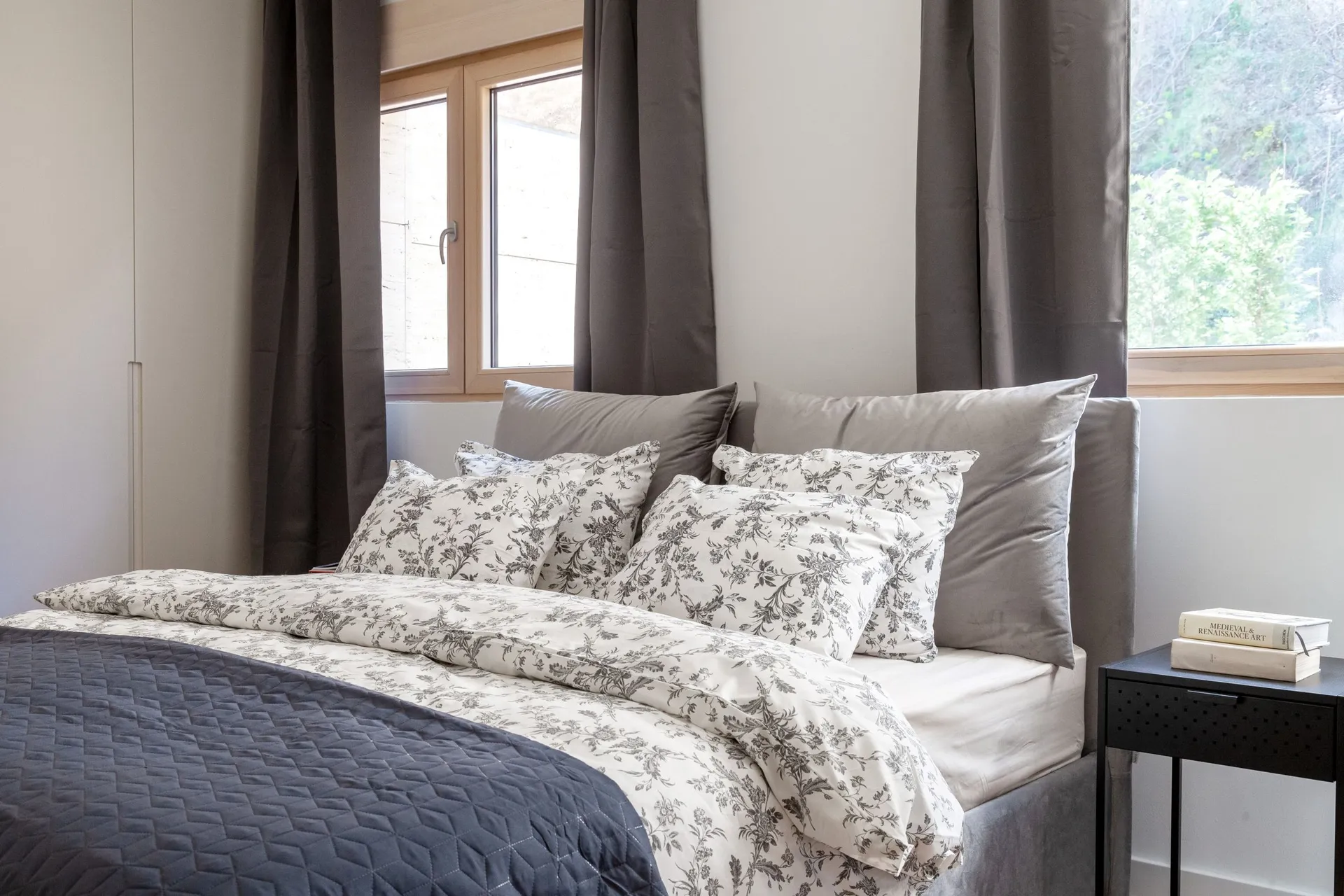 Modern bedroom featuring a double bed with a grey headboard and floral patterned bedding near windows.