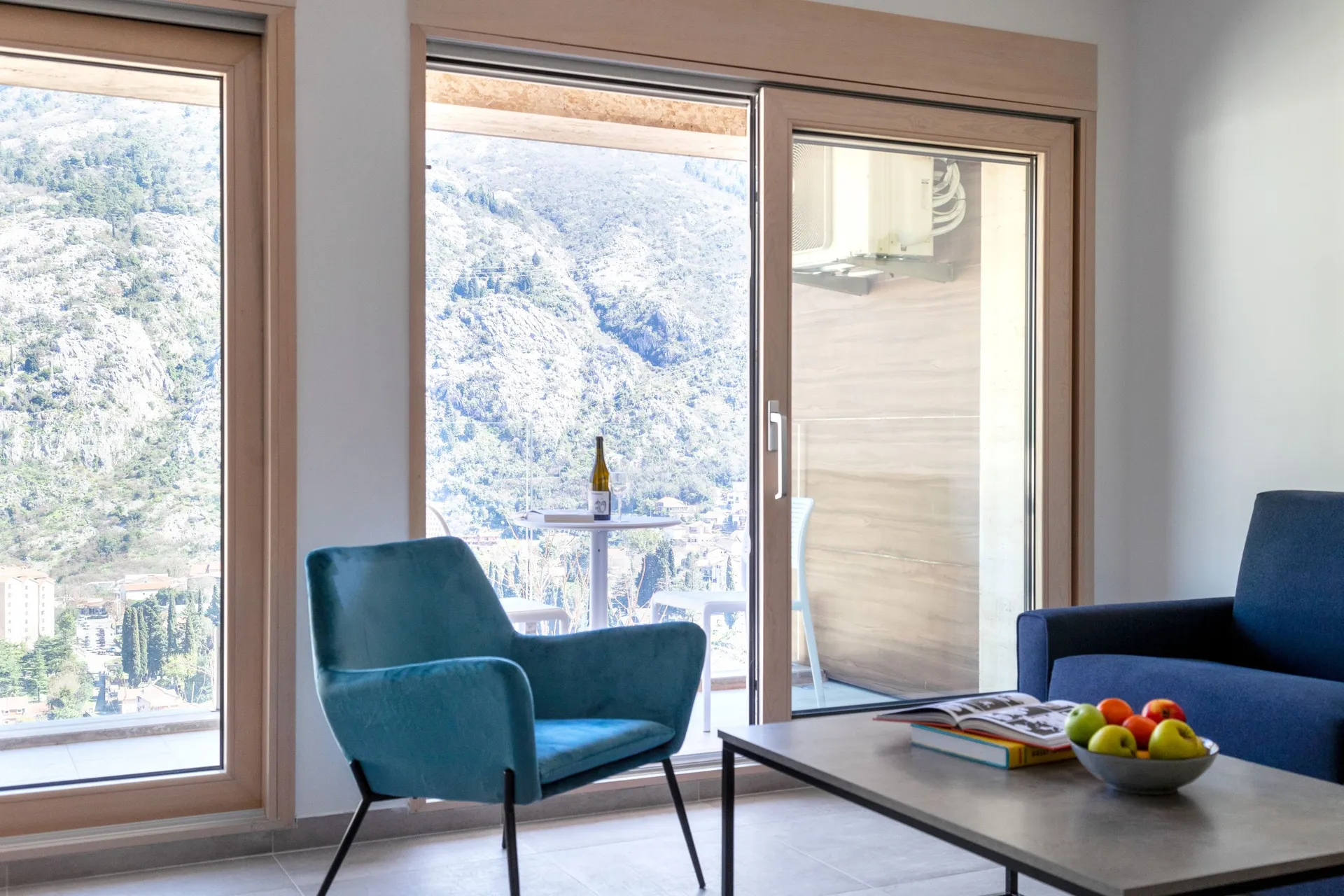 Bright living room with a turquoise chair and blue sofa. A table holds fruit and books. A mountain view is visible through the window.
