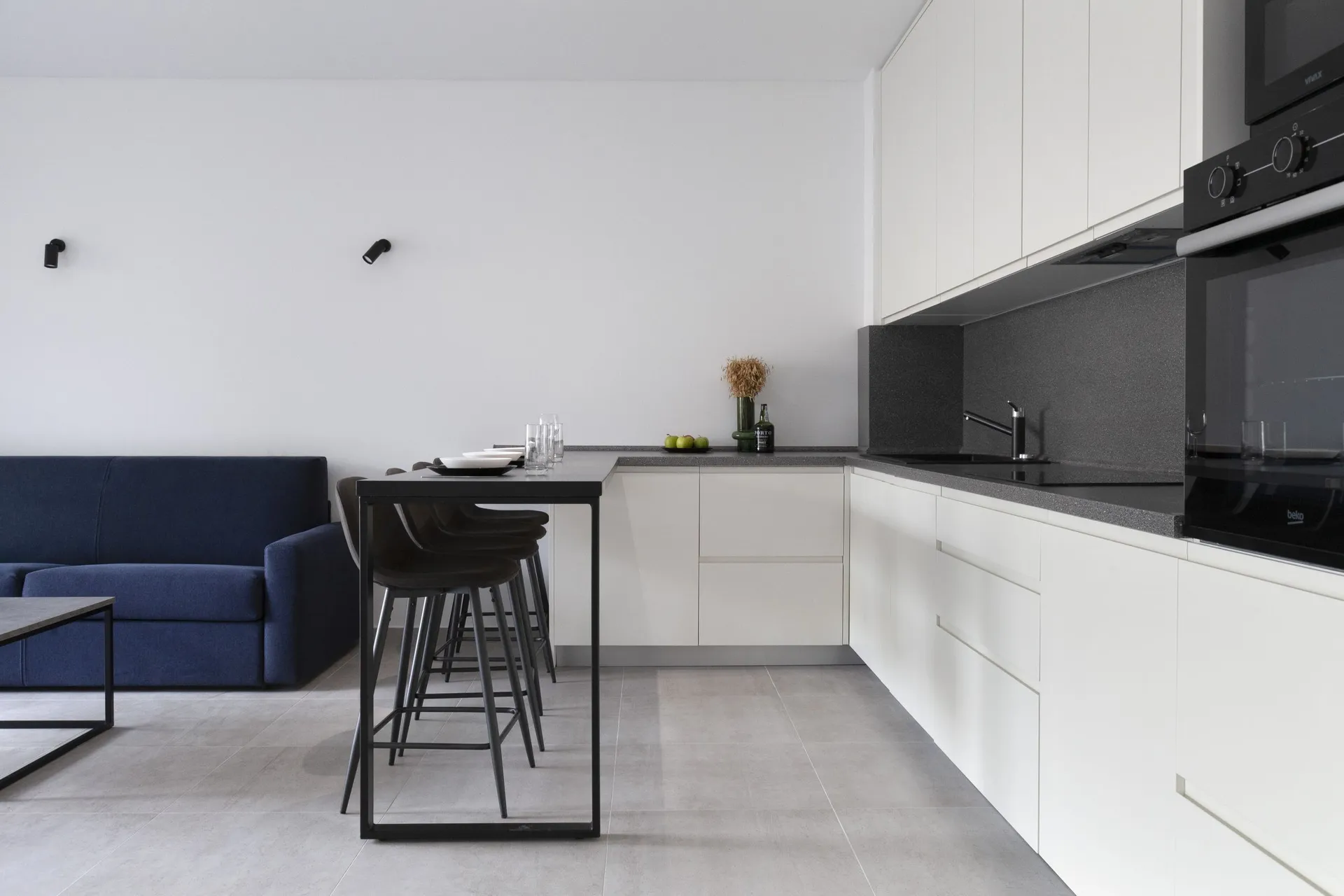Modern kitchen with white cabinets, black countertop, and bar stools next to a blue sofa in an open plan layout.