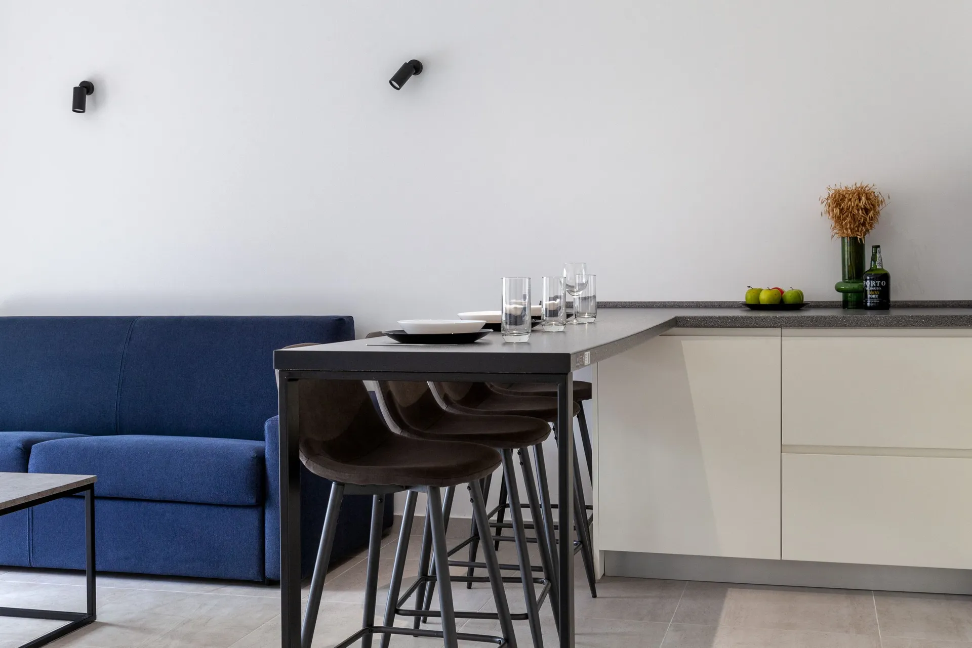 Modern open-plan living area with a blue sofa, black bar table, and white kitchen cabinets.