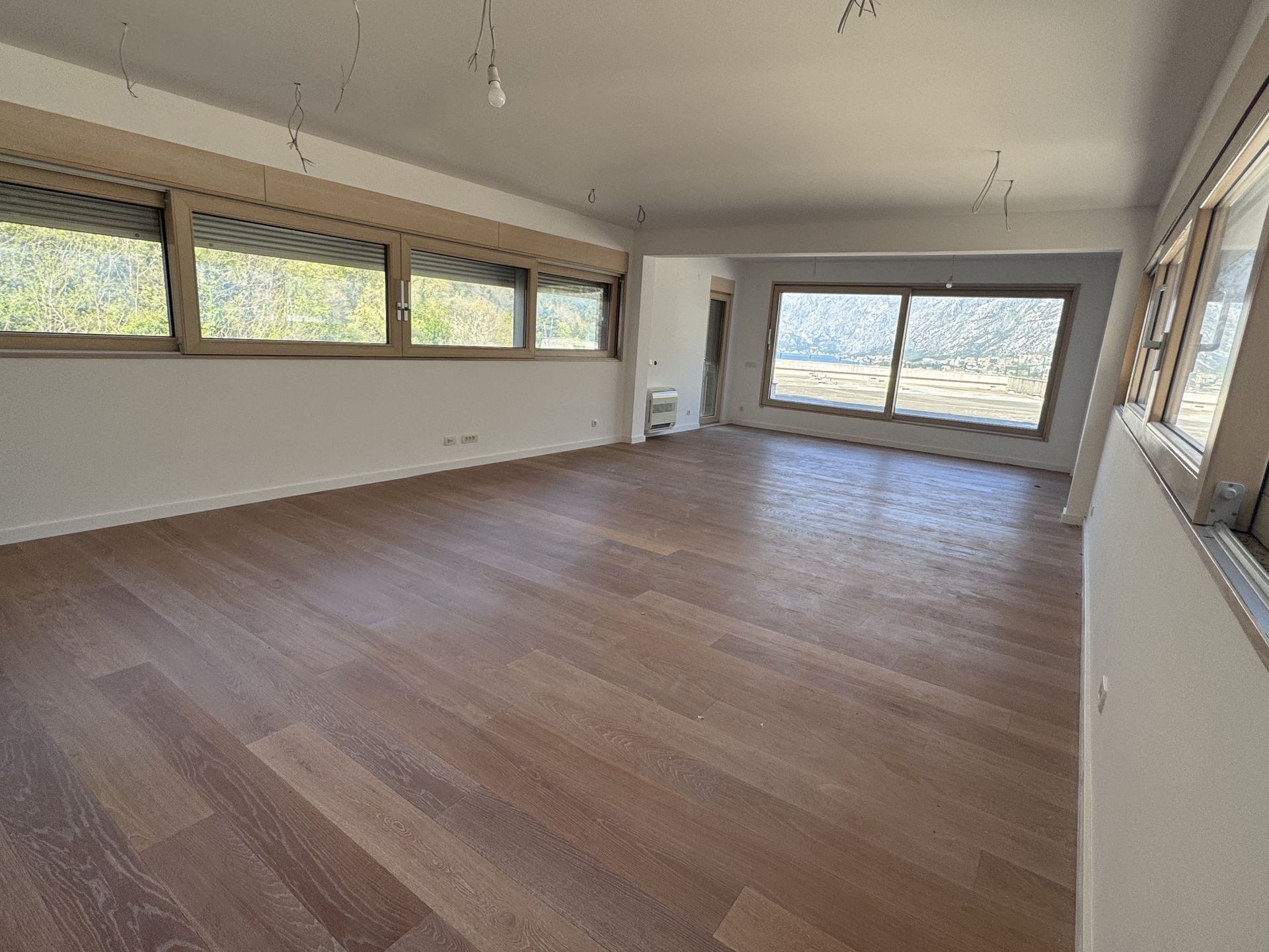 Spacious living room in a duplex in Kotor, Montenegro: panoramic windows, mountain view, and wooden flooring, unfurnished.