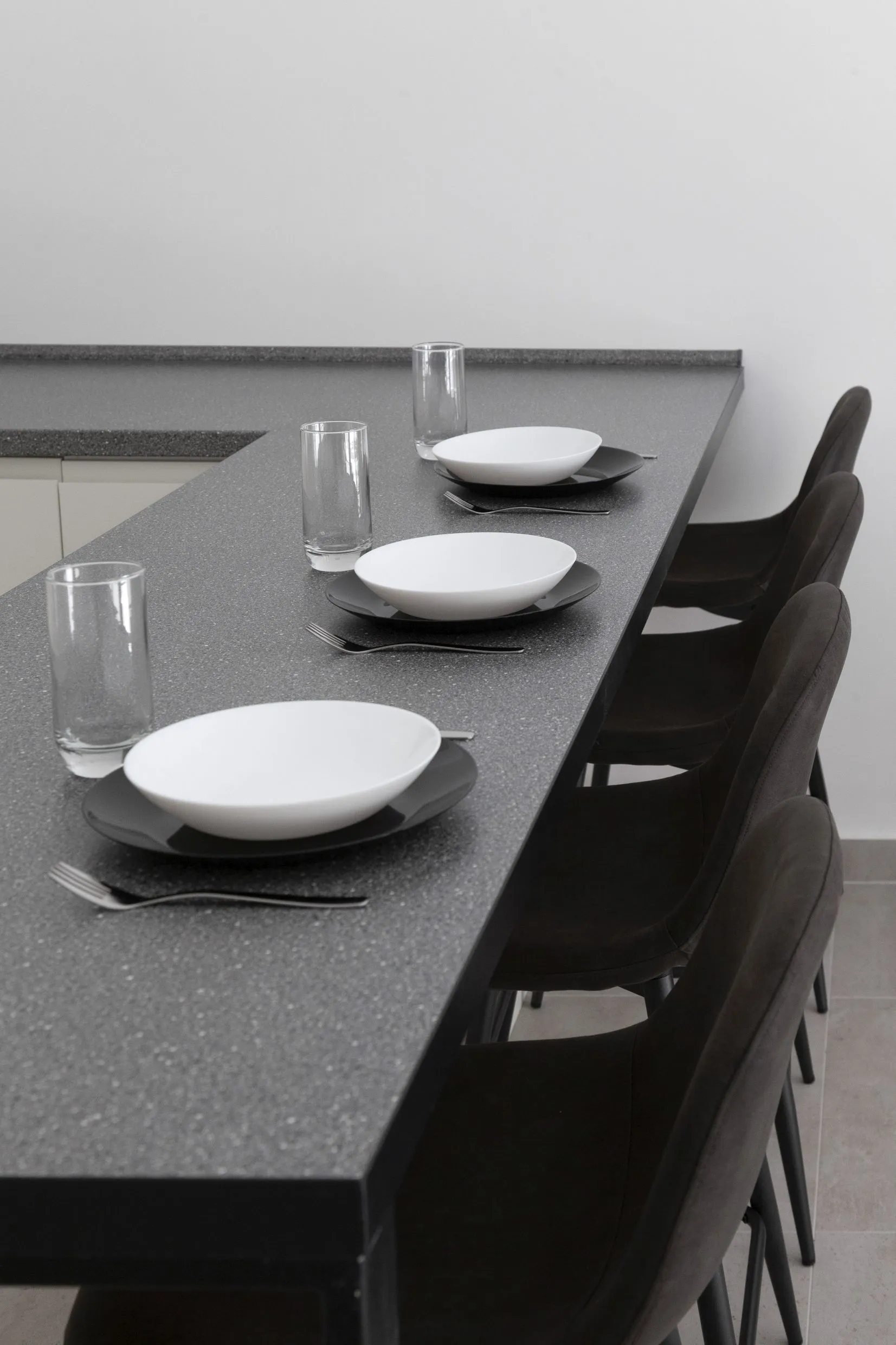 Modern kitchen counter with dark surface set with white bowls, glasses and forks alongside dark upholstered chairs against a white wall.