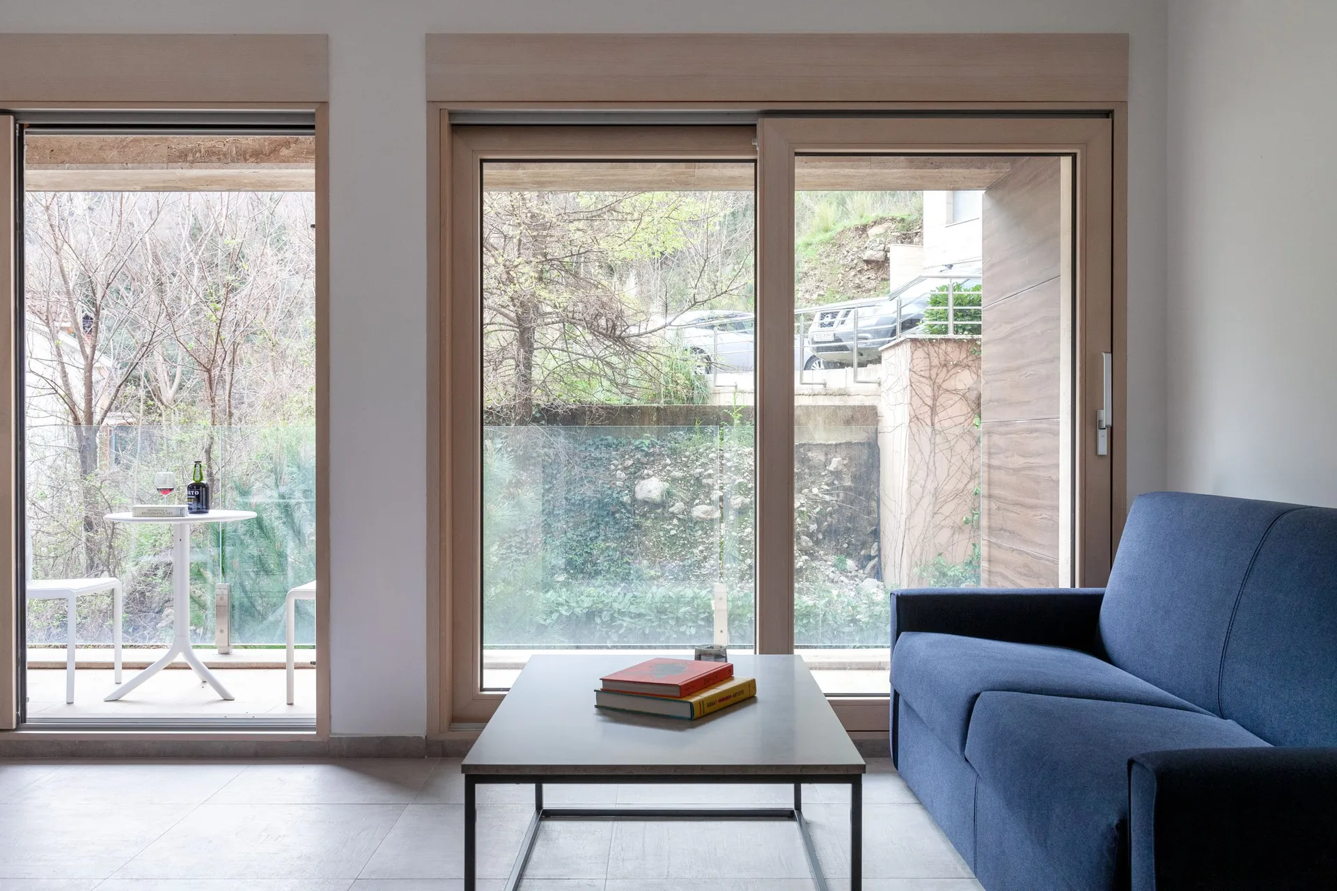 Modern living room with a blue sofa, coffee table, and books. Large windows lead to a balcony overlooking greenery and trees.
