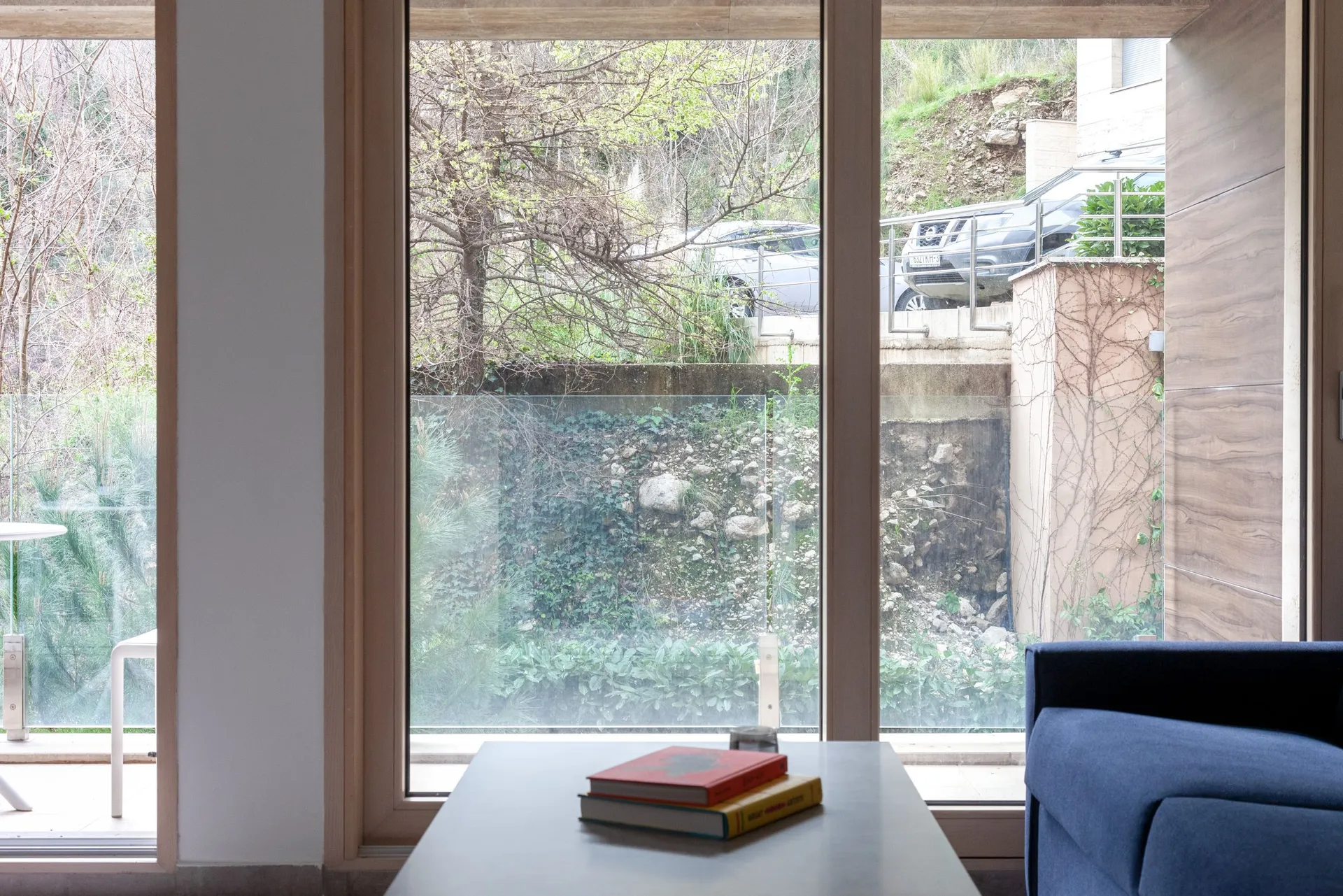 Modern living room with a blue sofa and table by the window. Books are on the table. Outside, a green slope and parked cars are visible.