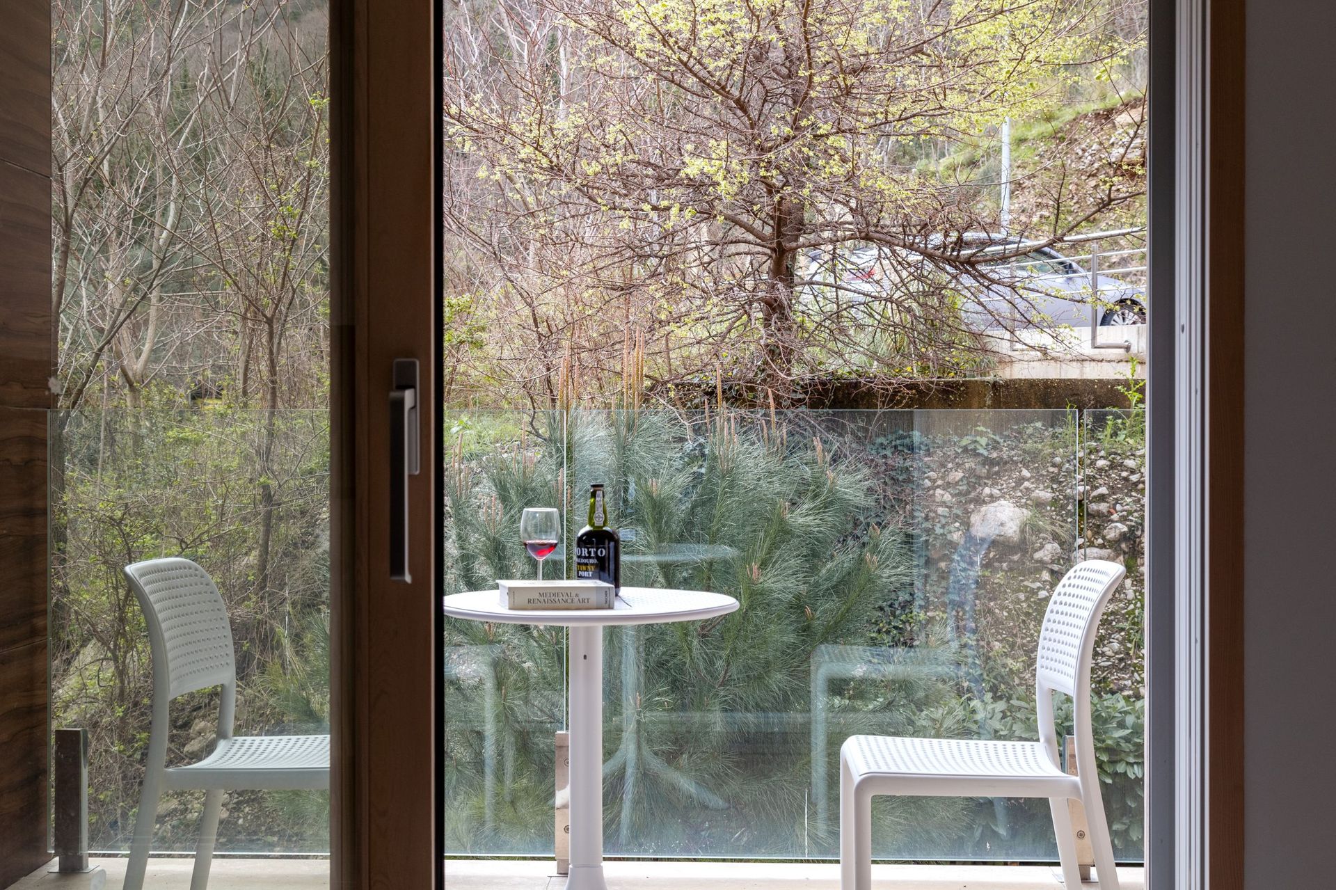 Apartment balcony in Kotor, Montenegro: a table with wine and a view of a green garden through a glass railing.