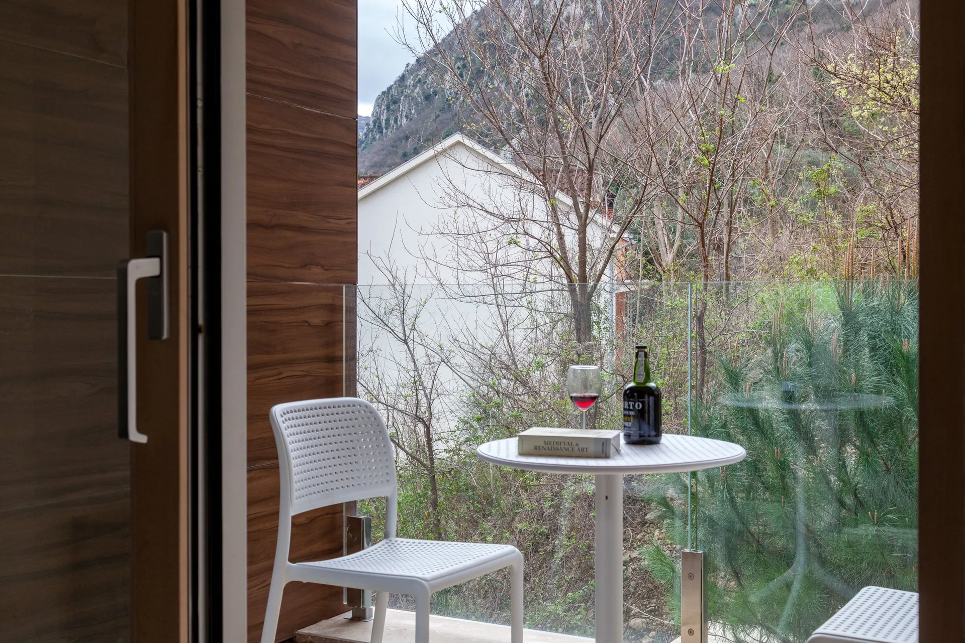 A white plastic chair and round table with a book, wine glass, and bottle on a balcony overlooking mountains and trees.