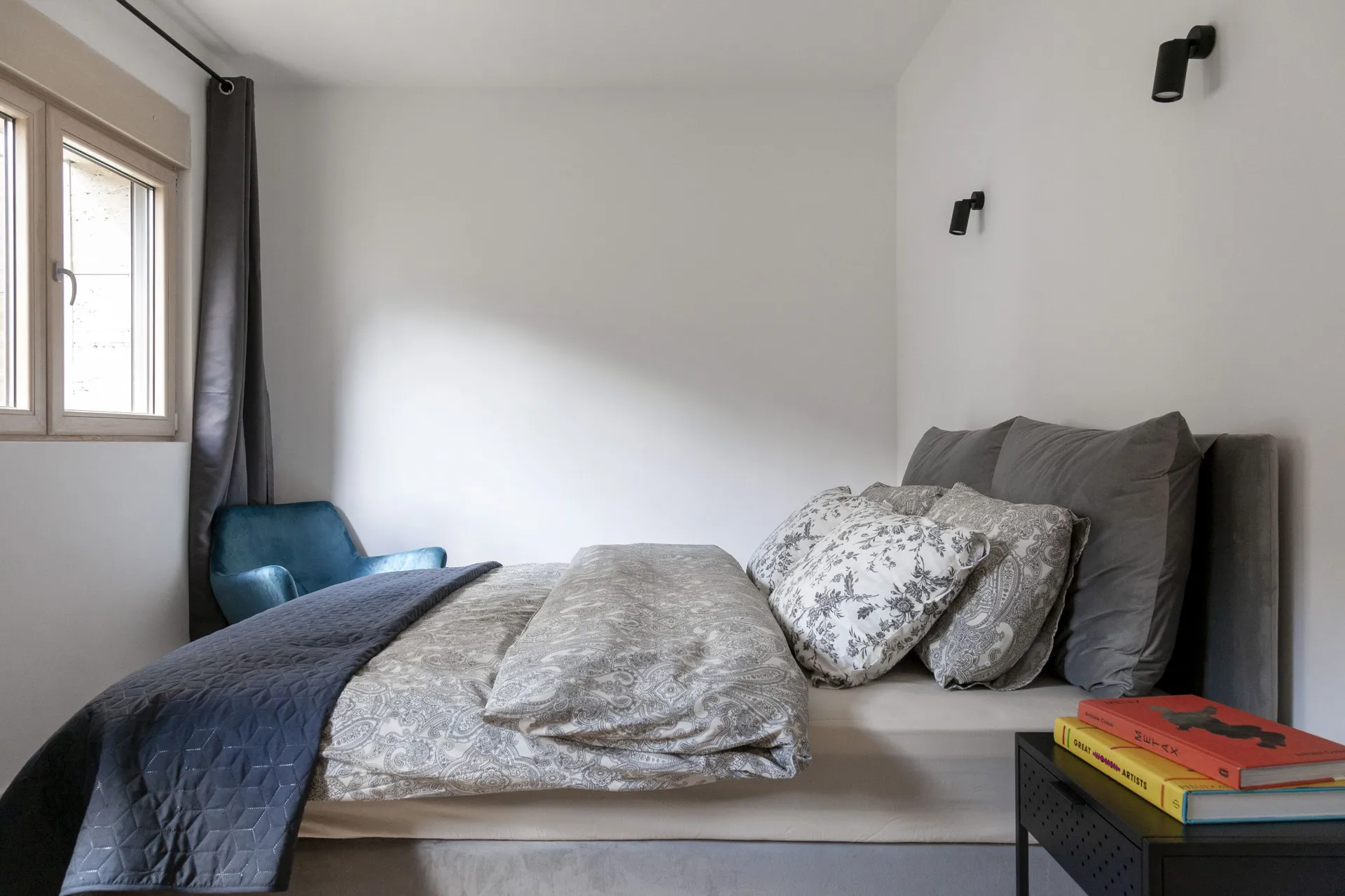 Modern bedroom featuring a gray bed with patterned bedding, a blue armchair by the window, and black wall sconces.