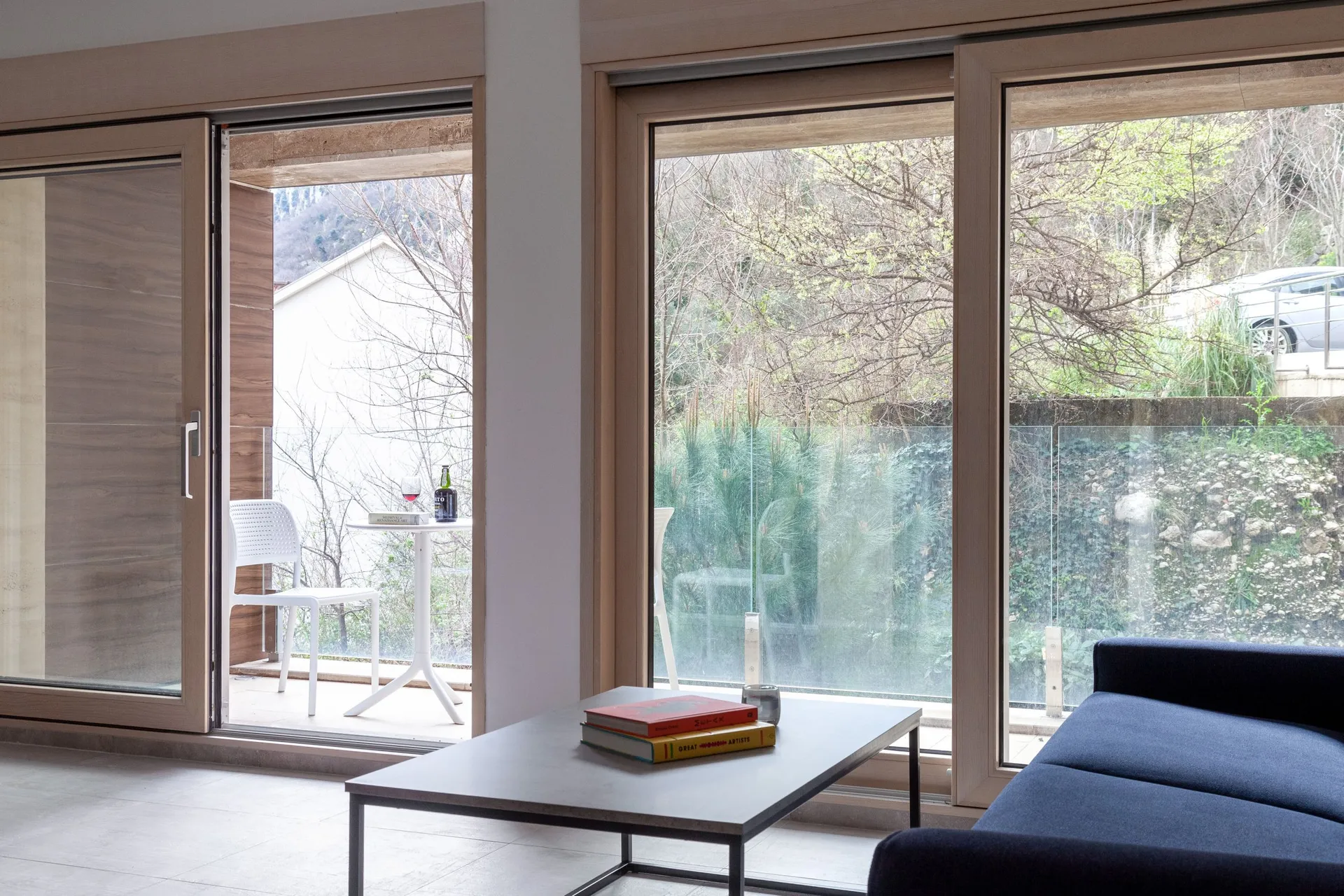 Modern living room with a blue sofa and grey table near panoramic windows. A balcony with white chairs and a view of nature is visible.