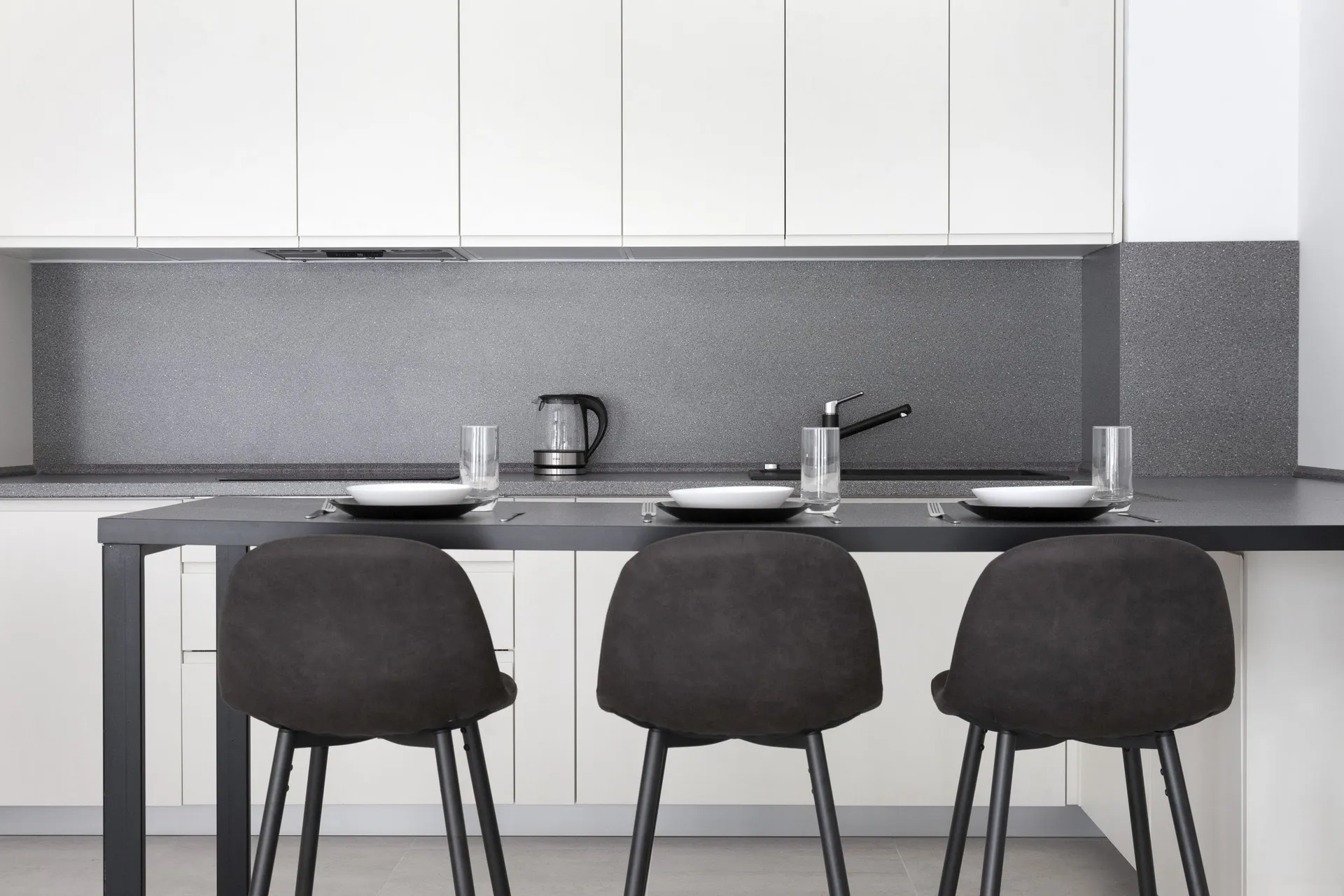 Modern kitchen with white cabinets, gray backsplash, and black bar counter featuring three stools and tableware.