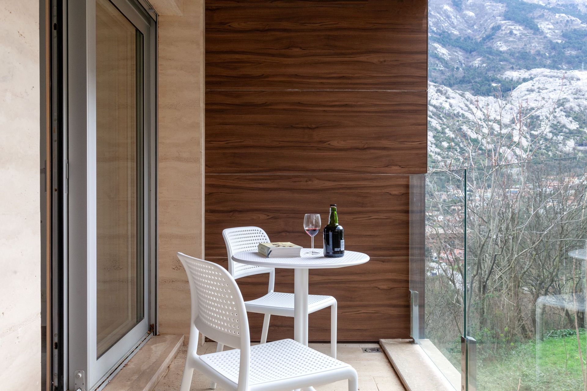 Apartment balcony in Kotor, Montenegro: white furniture, wood finish, glass railing, and mountain view.