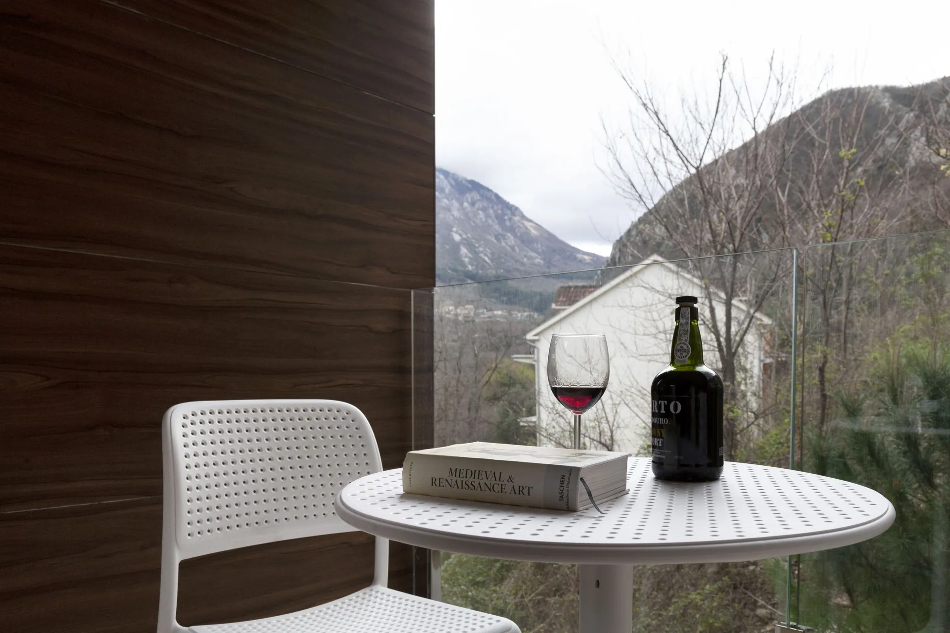 White perforated table and chair on a balcony overlooking mountains. A book, wine bottle, and glass sit on the table.