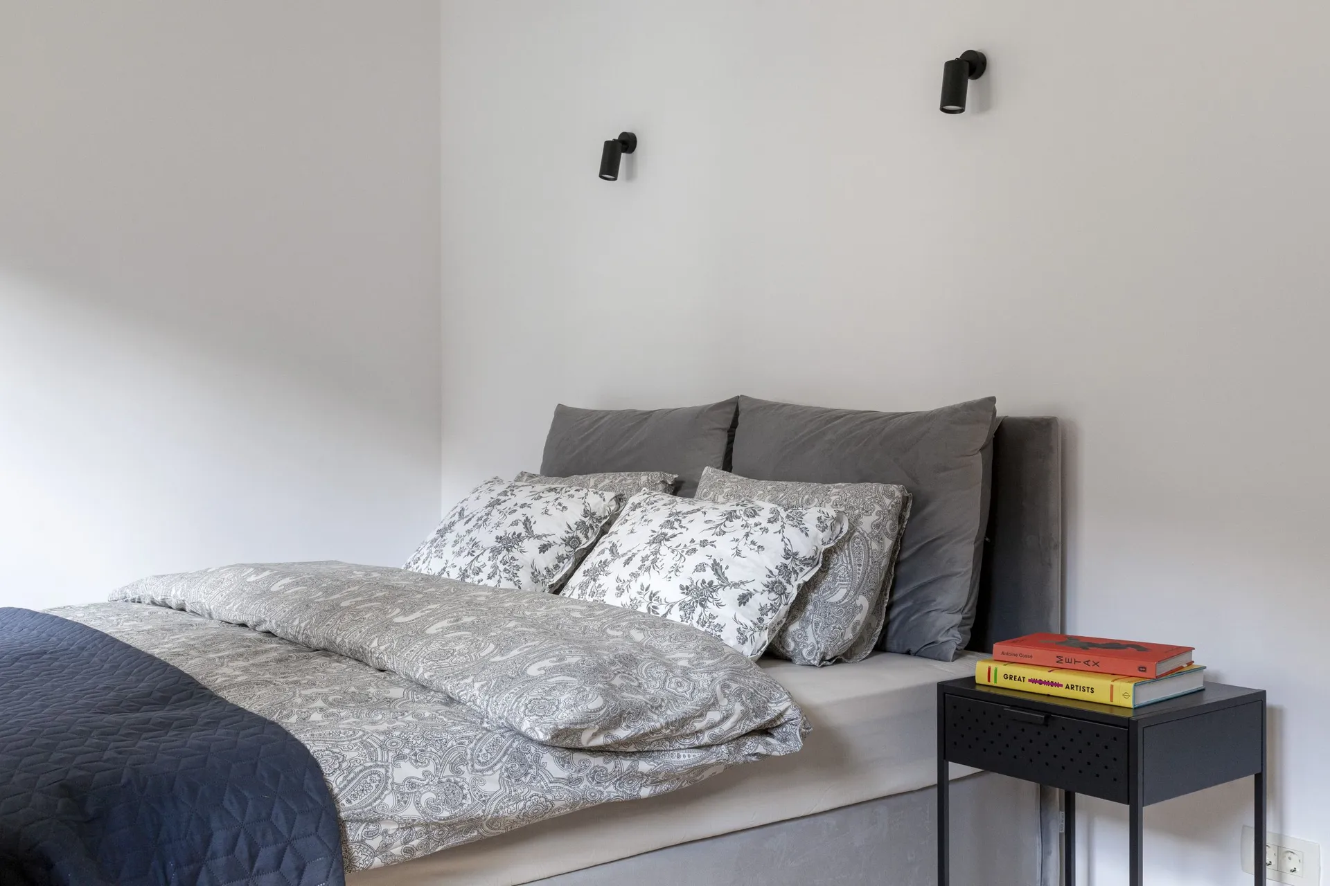Modern bedroom featuring a gray upholstered bed, patterned bedding, and black wall-mounted spotlights positioned above the headboard.