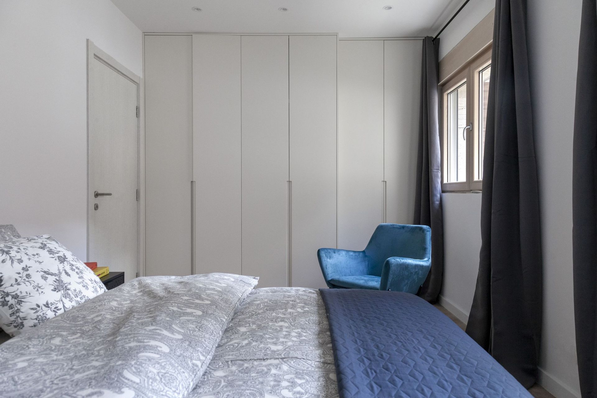 Modern bedroom in an apartment in Kotor, Montenegro: built-in wardrobe, patterned bed, and a blue armchair by the window.
