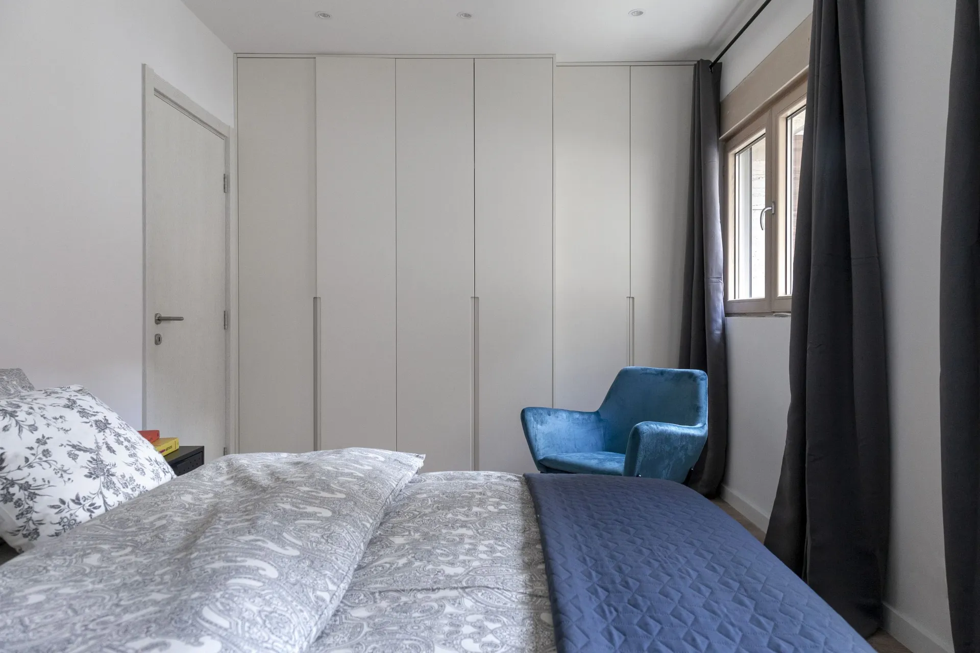 Modern bedroom featuring a large white built-in wardrobe, a blue velvet armchair, and a bed dressed in grey and blue patterned bedding.