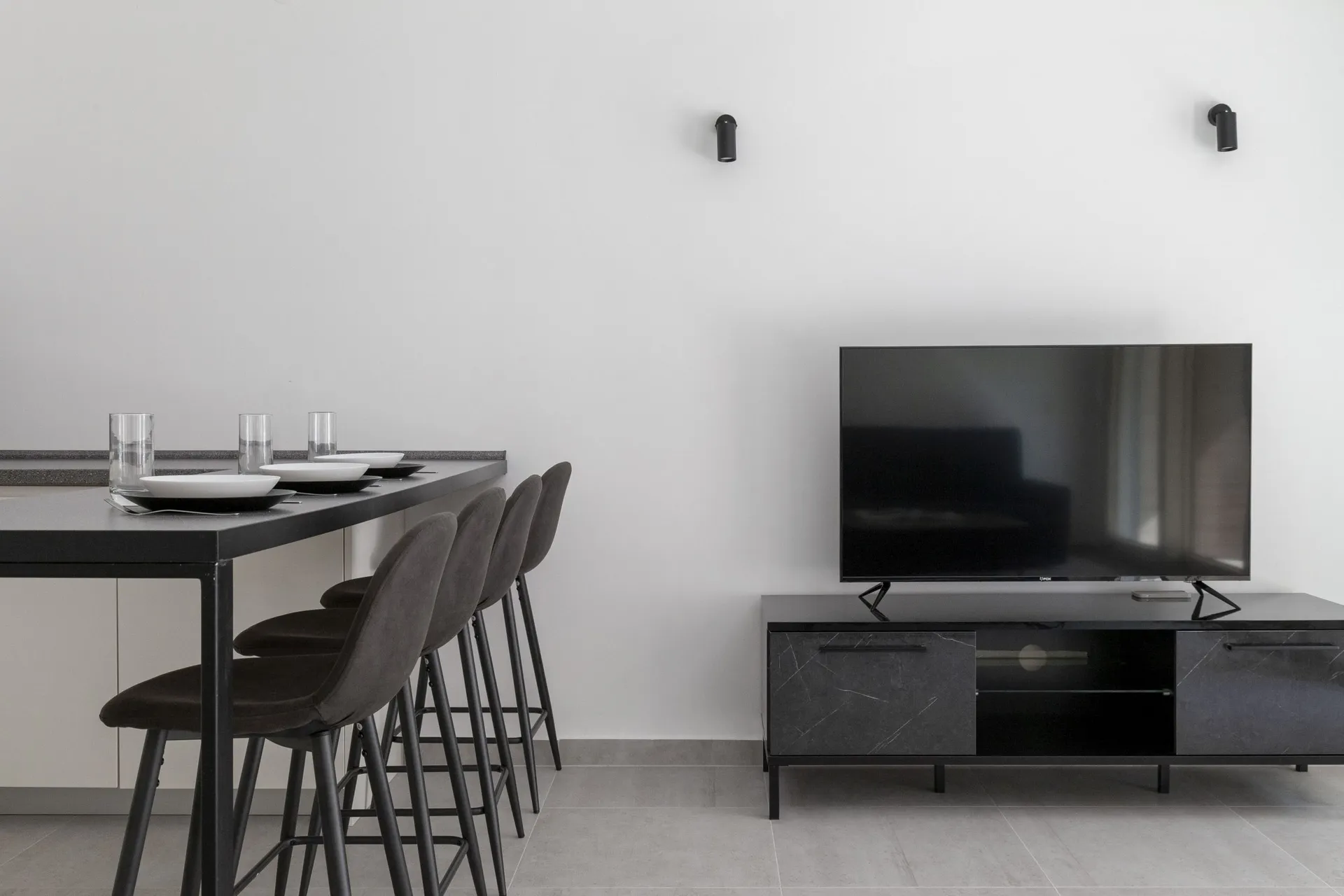 Modern living area with a black bar counter, gray stools, and a TV on a dark console against a white wall.